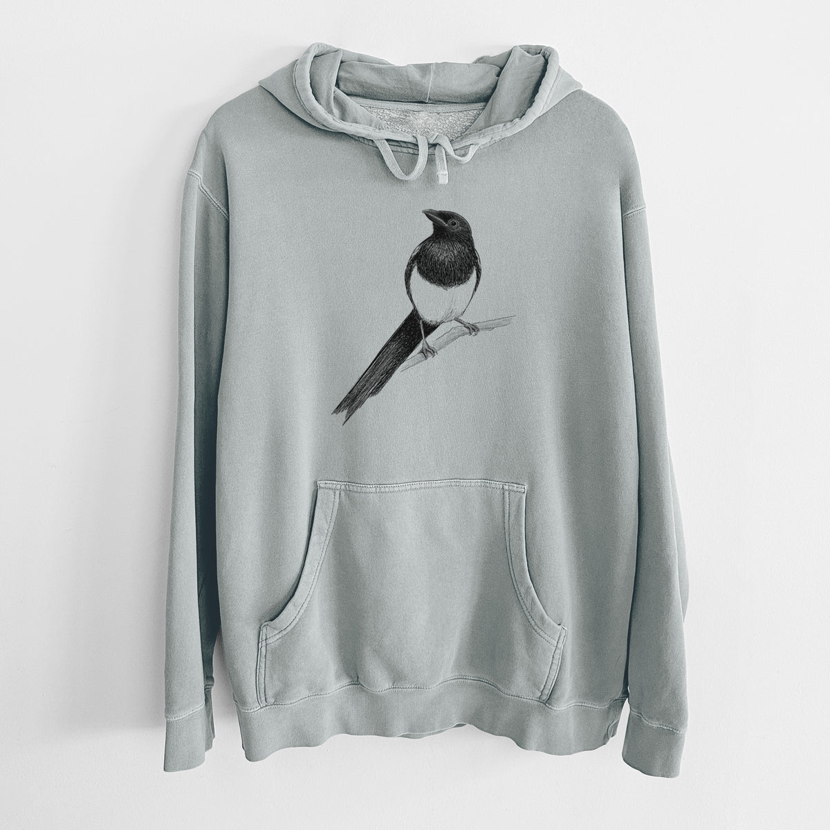Sage green pigment dyed hoodie laid flat, printed with a detailed black ink sketch of a black-billed magpie perched on a branch with its long tail.