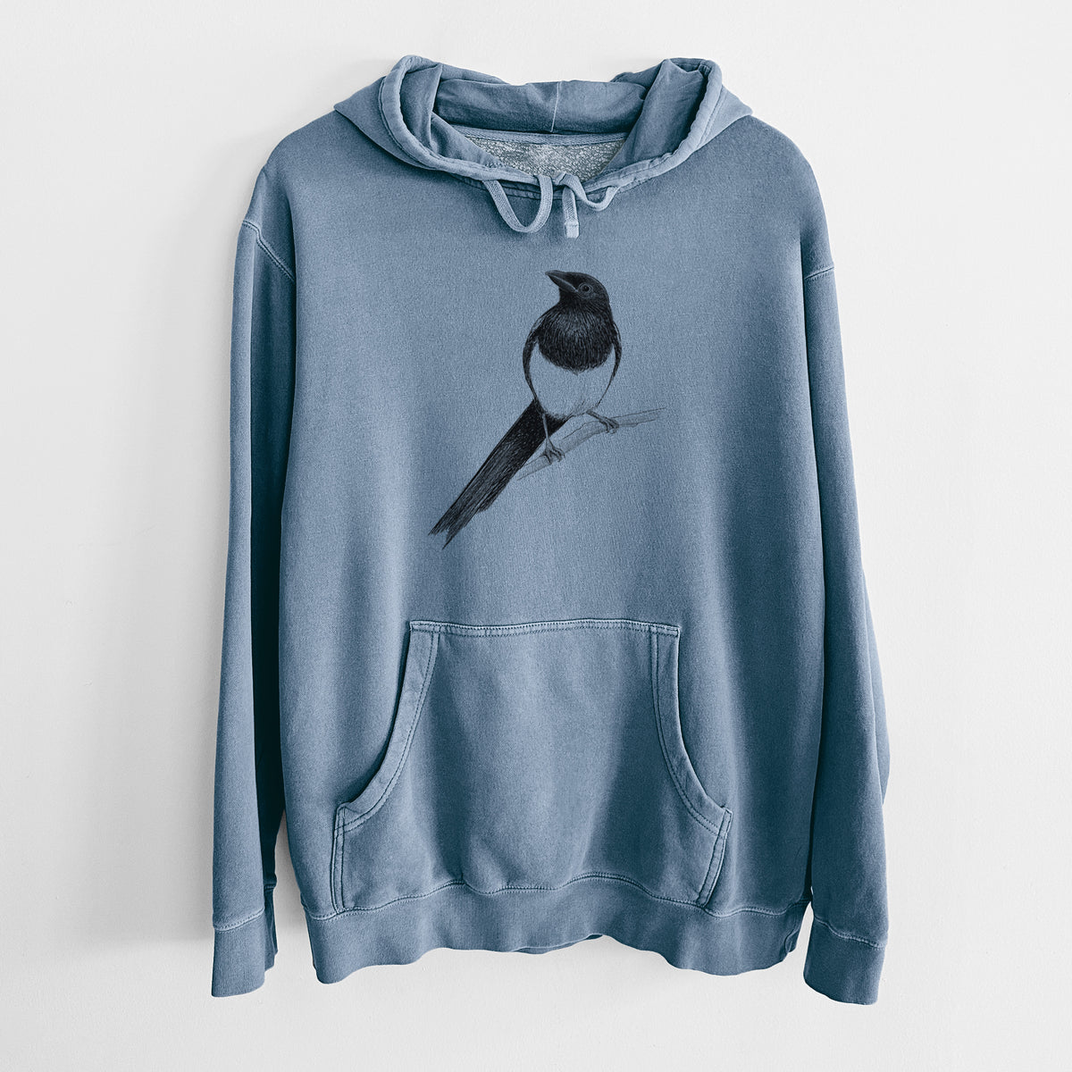 Slate blue pigment dyed hoodie laid flat, printed with a detailed black ink sketch of a black-billed magpie perched on a branch with its long tail.