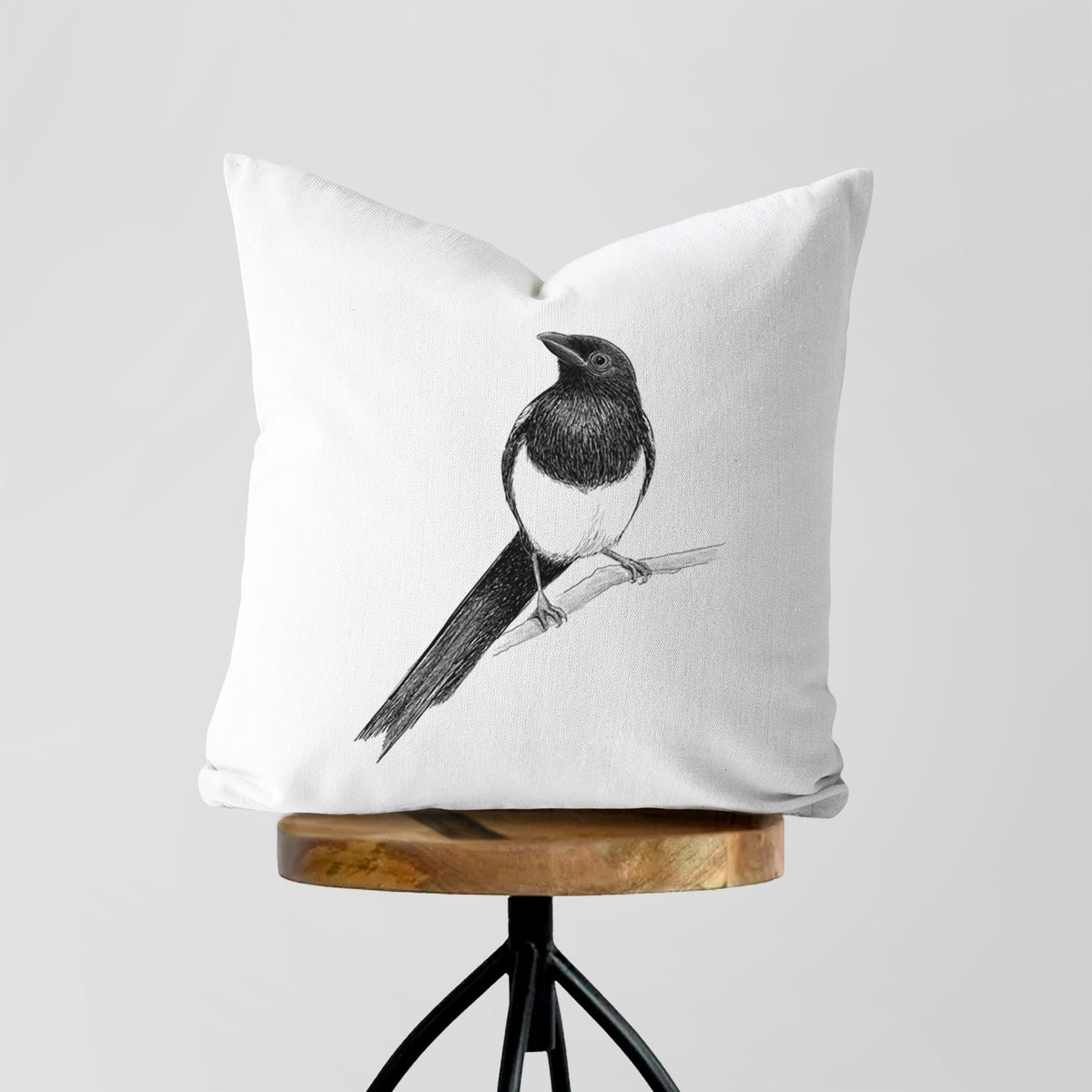White pillow cover printed with a detailed black ink sketch of a black-billed magpie perched on a branch with its long tail, on a wooden stool.
