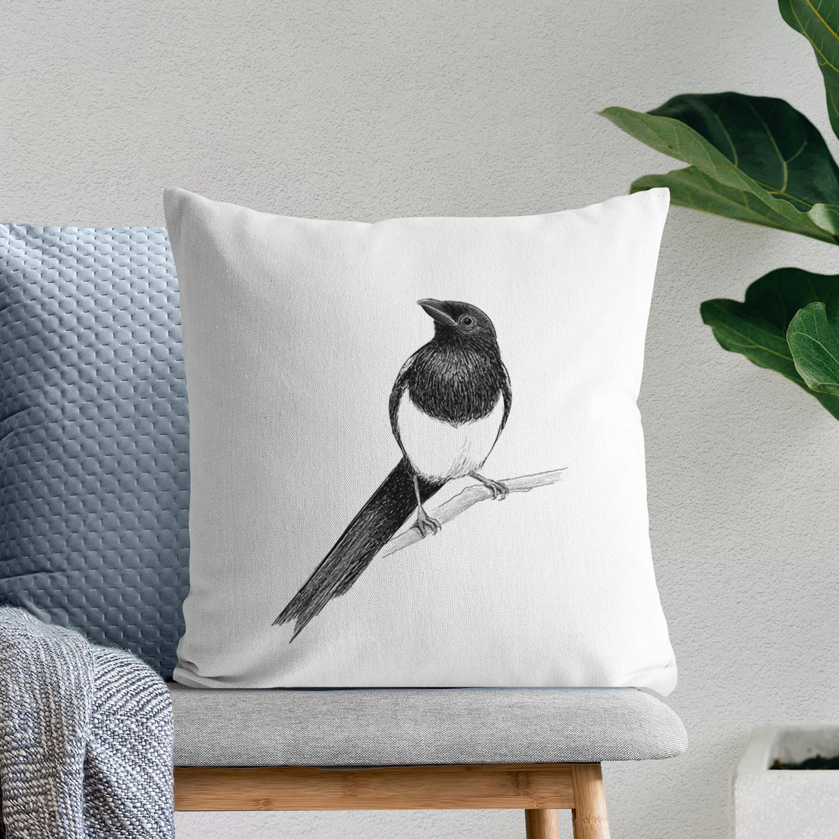 White pillow cover printed with a detailed black ink sketch of a black-billed magpie perched on a branch with its long tail, on a bench beside plants.