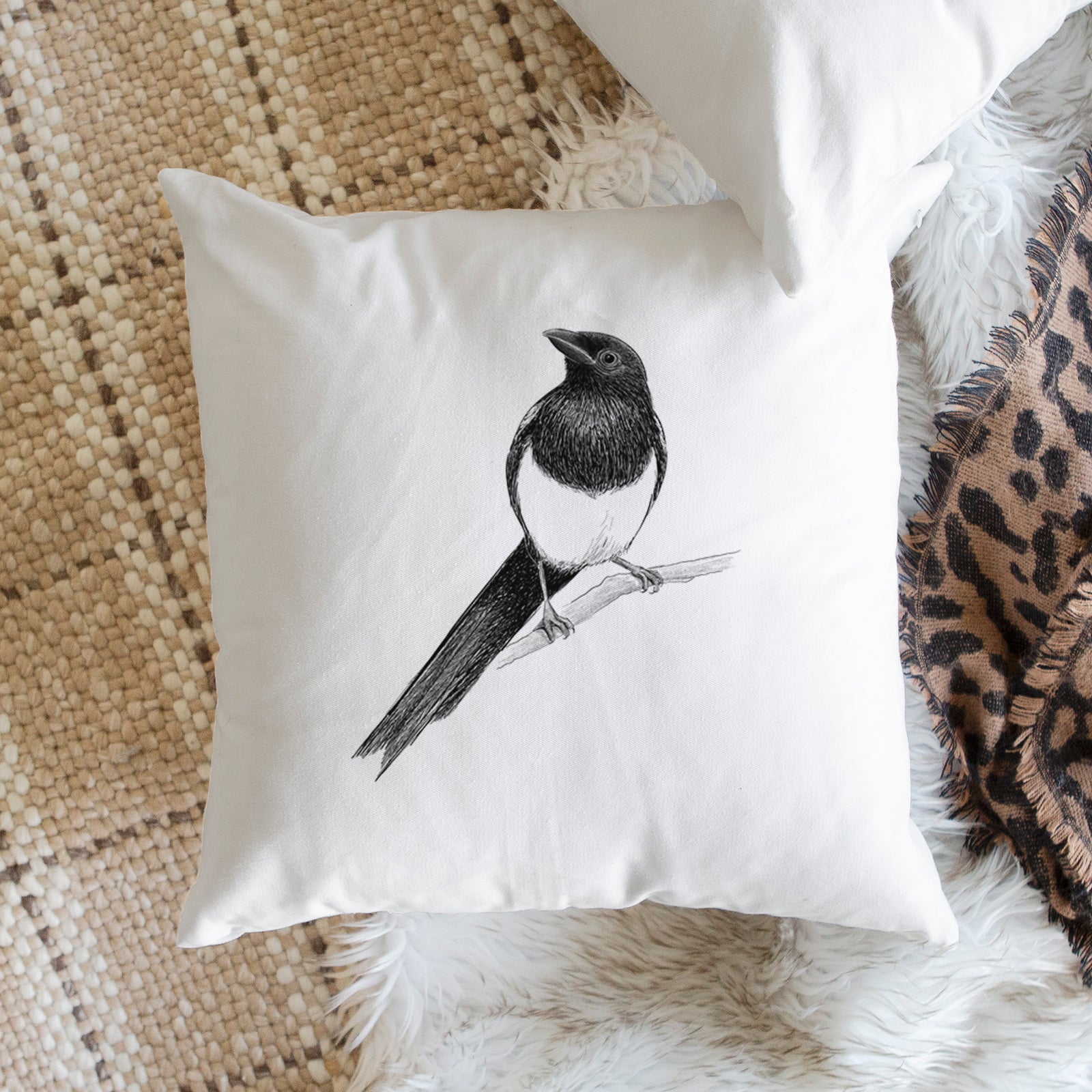 White pillow cover printed with a detailed black ink sketch of a black-billed magpie perched on a branch with its long tail, on a bench beside plants.