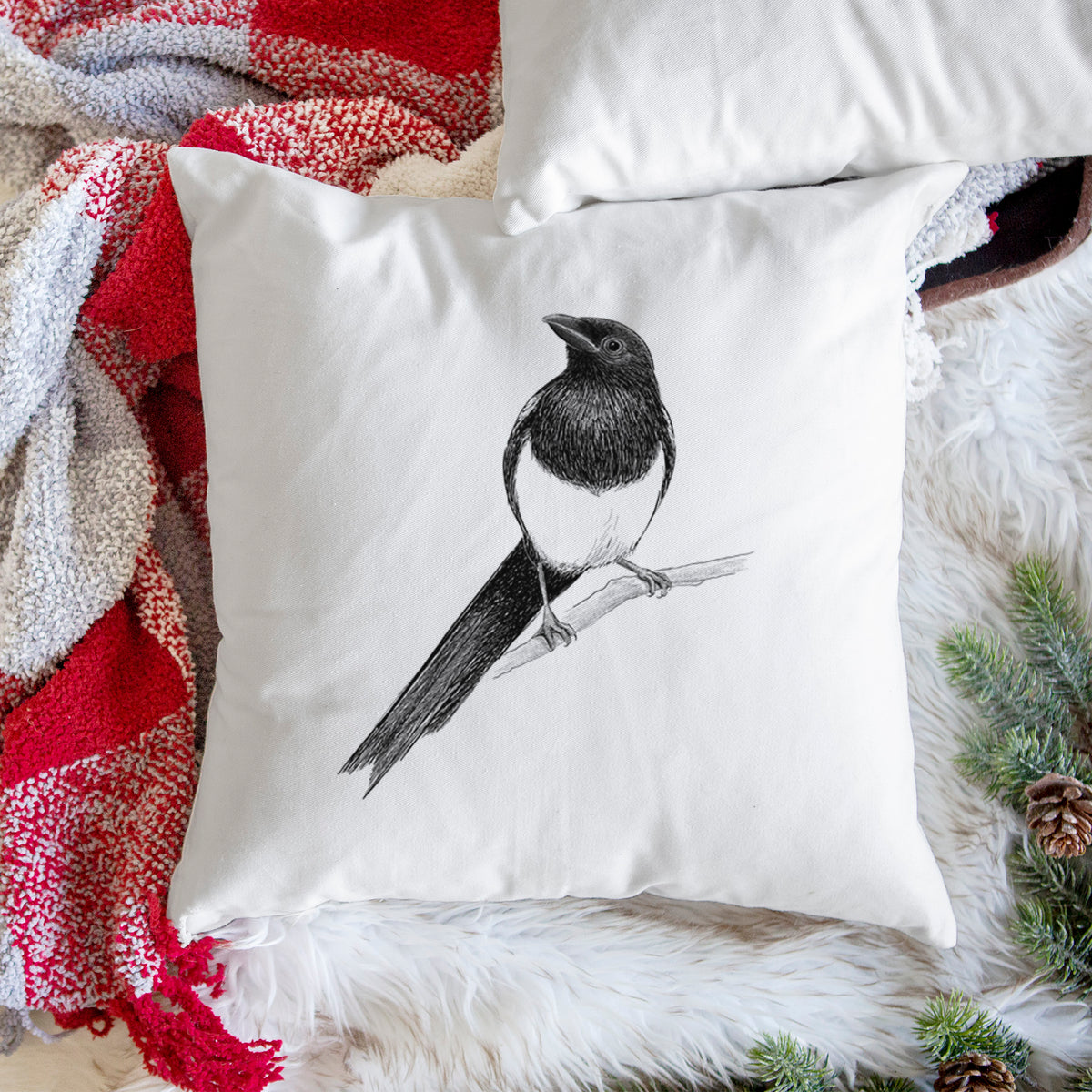 White pillow cover printed with a detailed black ink sketch of a black-billed magpie perched on a branch with its long tail, on a woven chair.