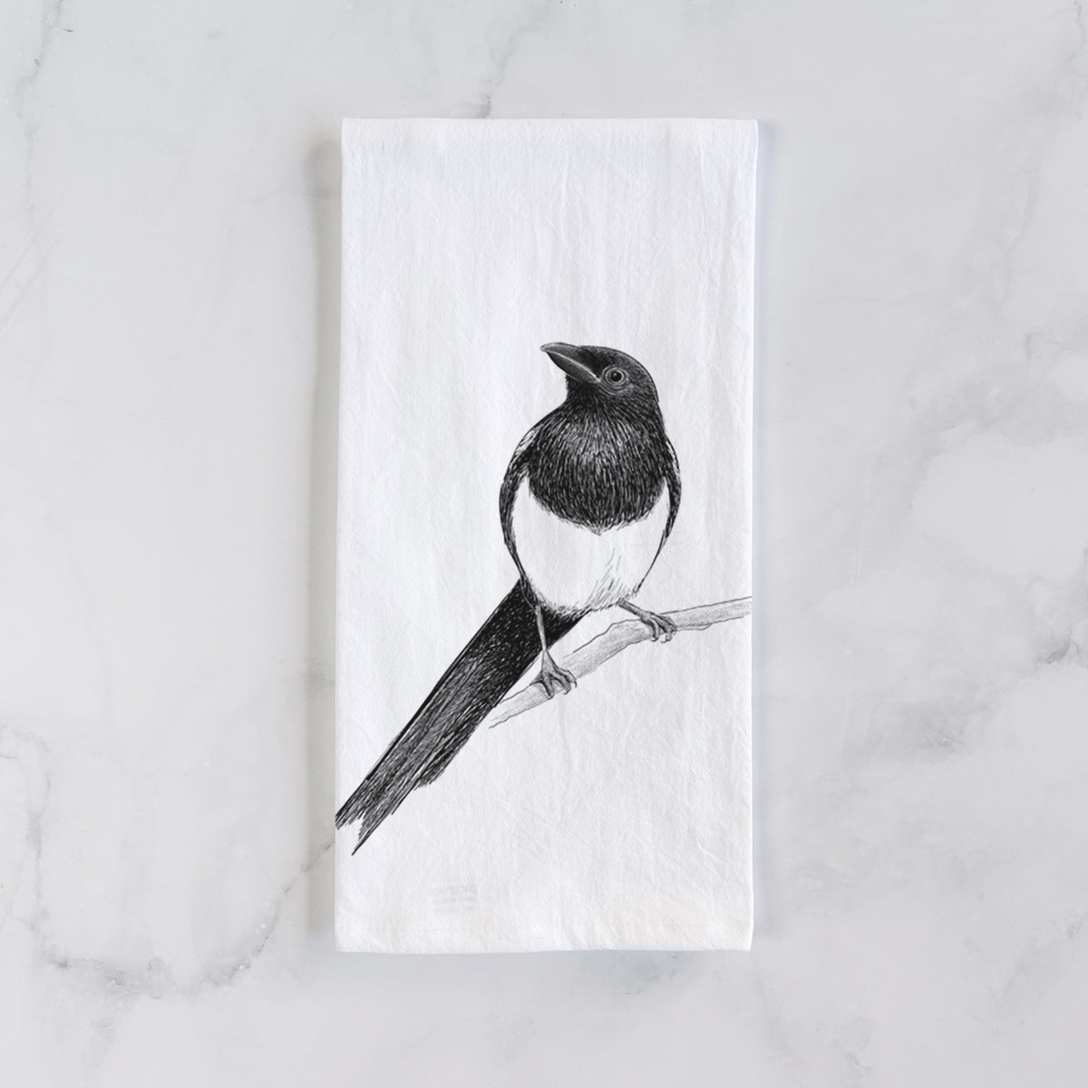 White cotton tea towel laid flat on marble, printed with a detailed black ink sketch of a black-billed magpie perched on a branch with its long tail.