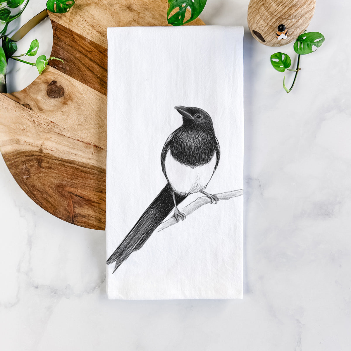 White cotton tea towel printed with a detailed black ink sketch of a black-billed magpie perched on a branch with its long tail, on a marble counter beside a round wooden cutting board and greenery.