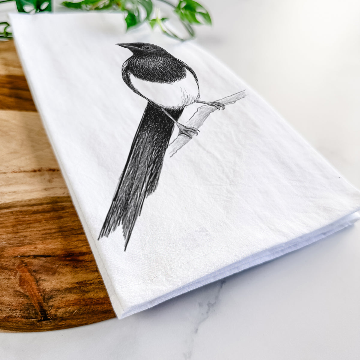 Close-up of a folded white tea towel on a wooden board, showing a detailed black ink sketch of a black-billed magpie perched on a branch with its long tail.