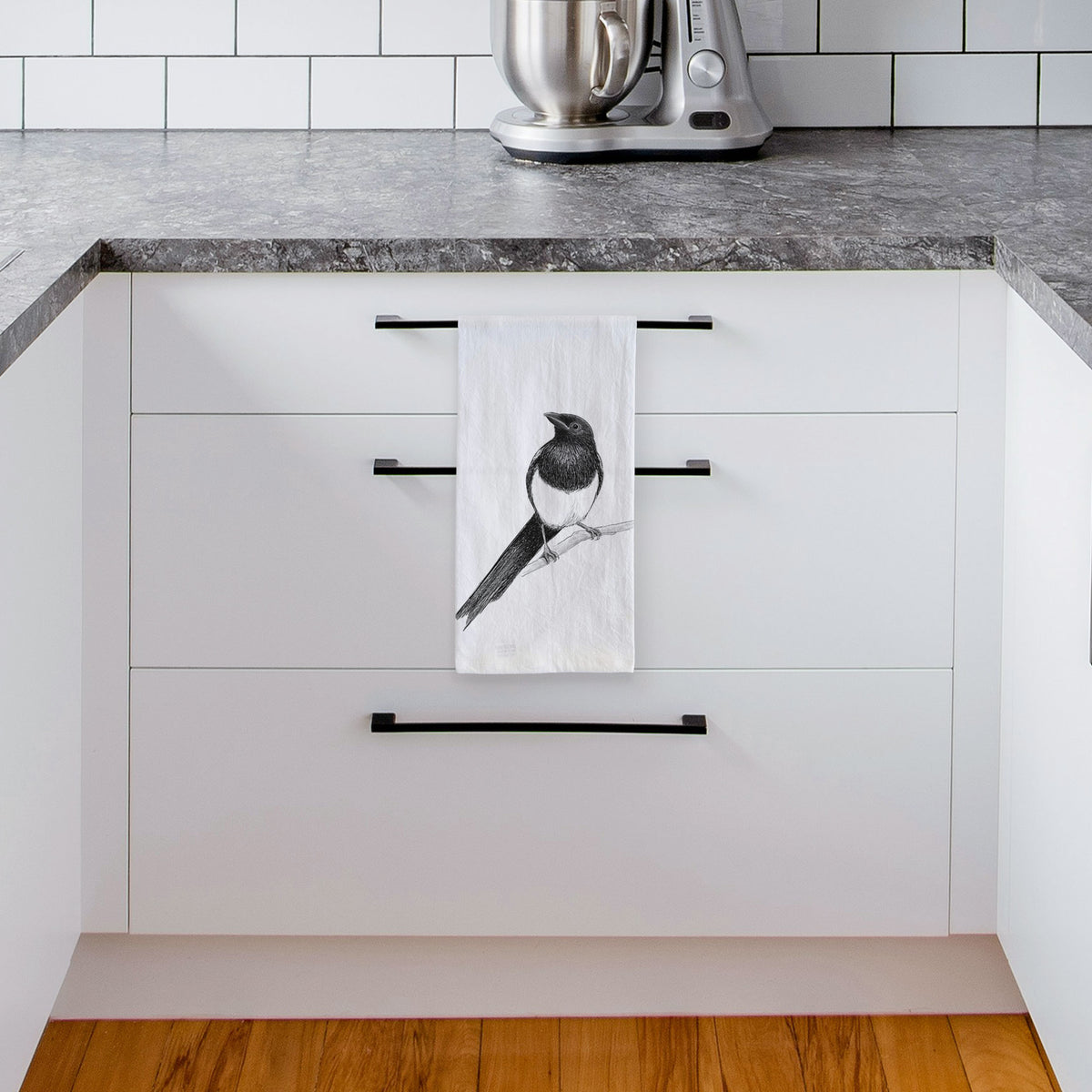 White tea towel printed with a detailed black ink sketch of a black-billed magpie perched on a branch with its long tail, hanging from a drawer handle in a bright white kitchen.