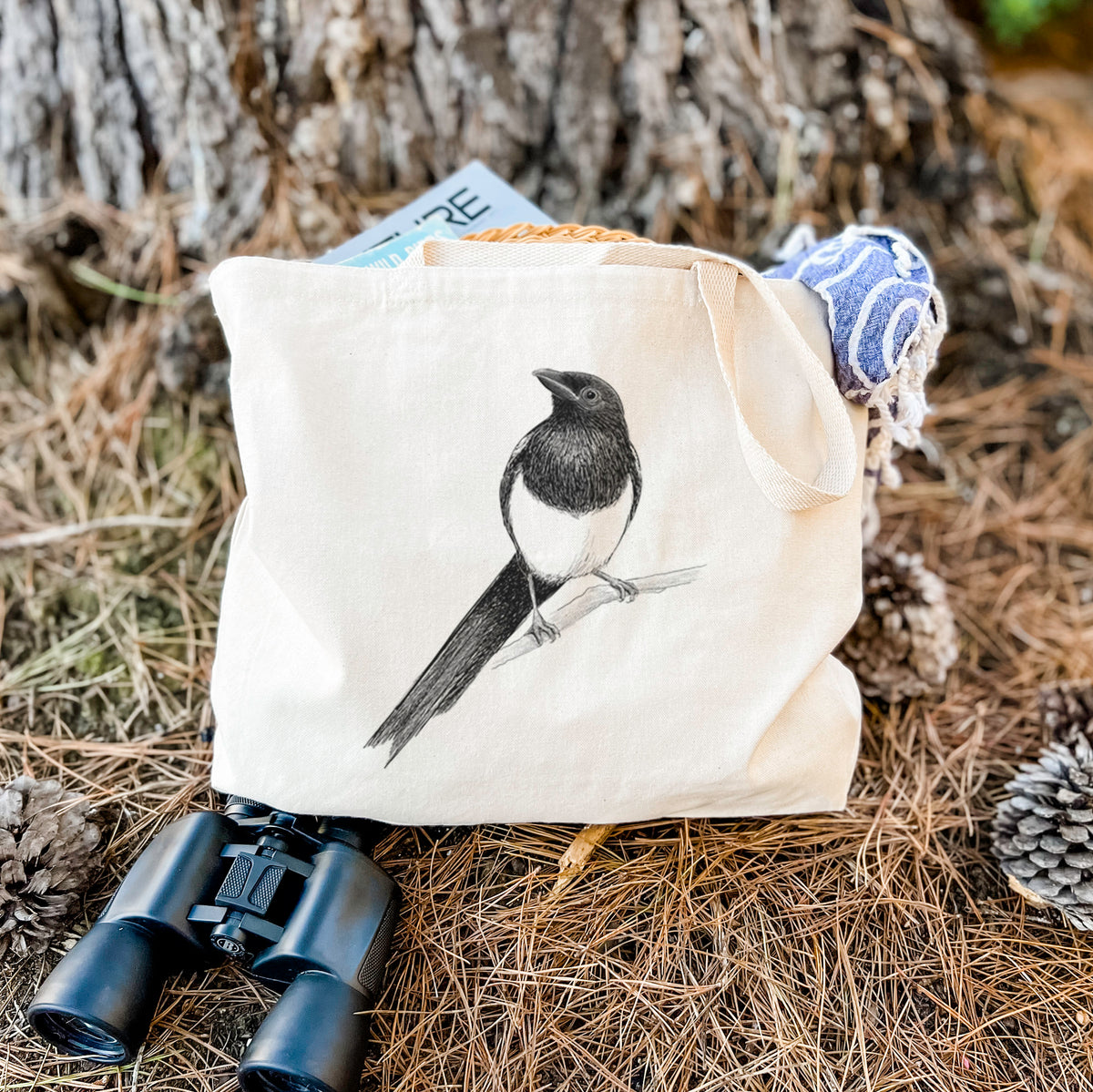 Canvas tote bag printed with a detailed black ink sketch of a black-billed magpie perched on a branch with its long tail, resting on dry grass beside a pair of binoculars.