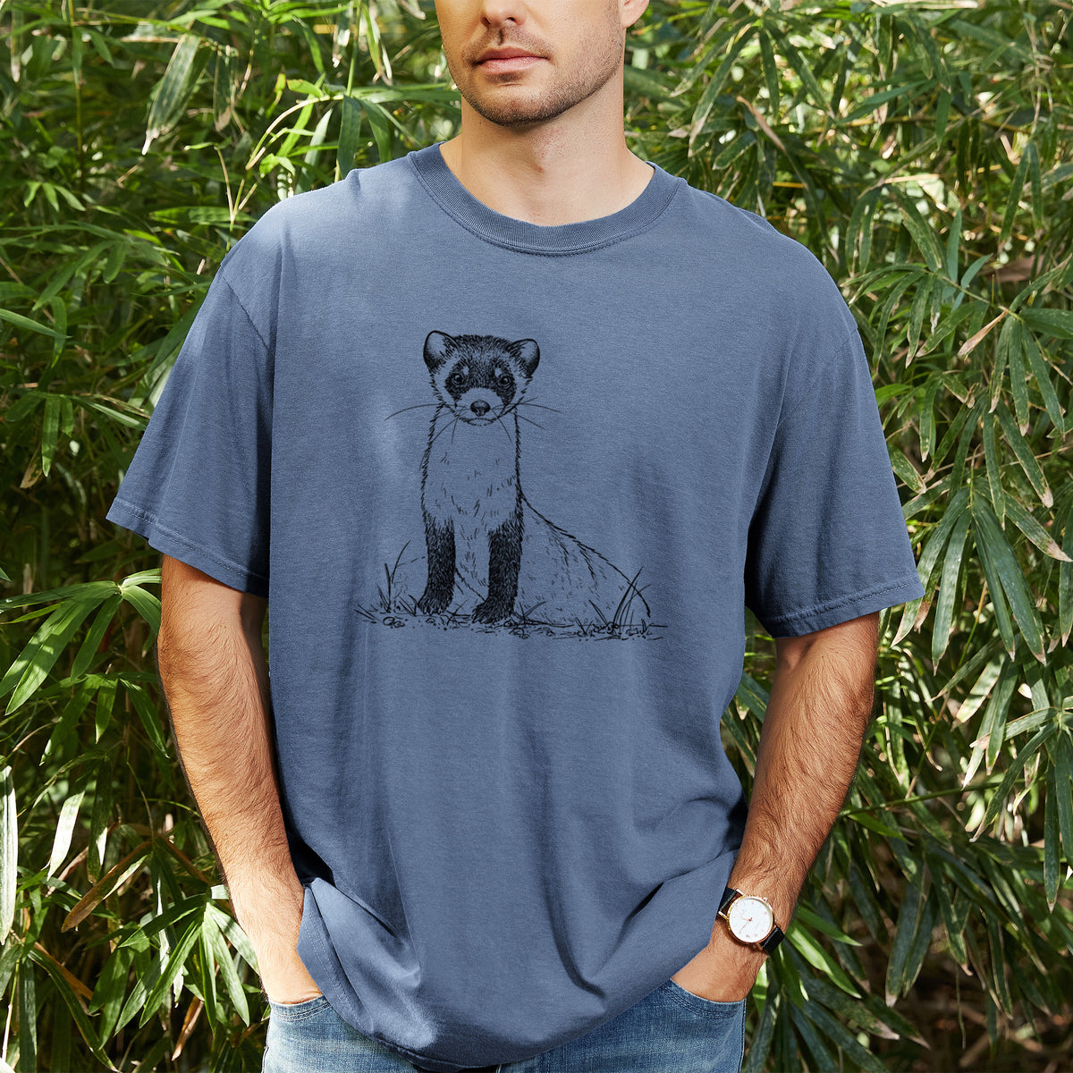 Man wearing a Comfort Colors t-shirt printed with a detailed black ink illustration of a black-footed ferret sitting on the ground, outdoors.