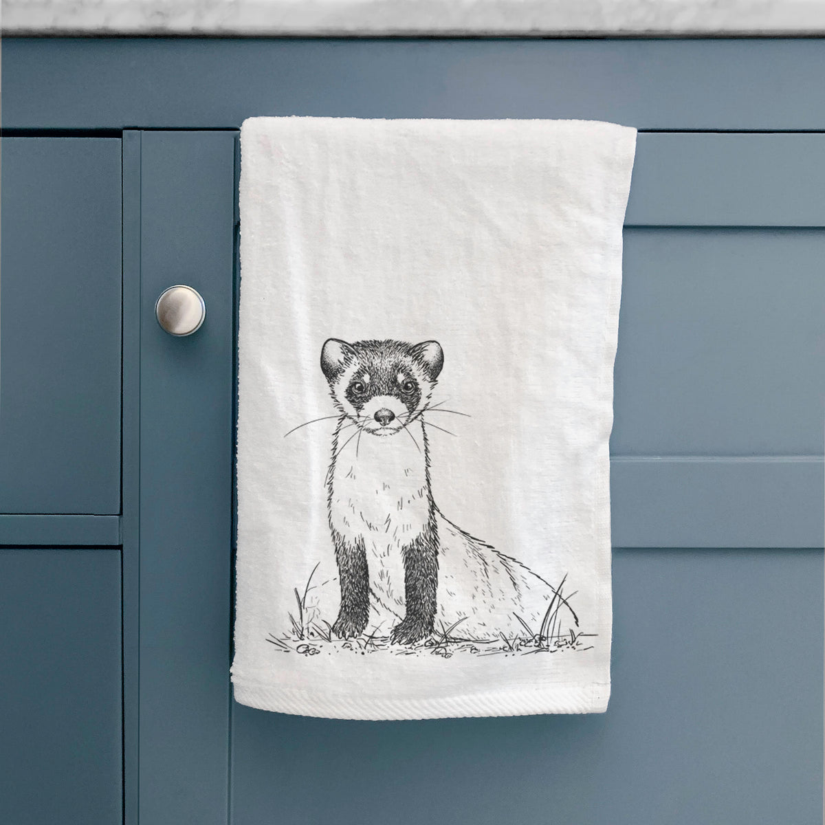 White hand towel printed with a detailed black ink illustration of a black-footed ferret sitting on the ground, hanging on a cabinet door.