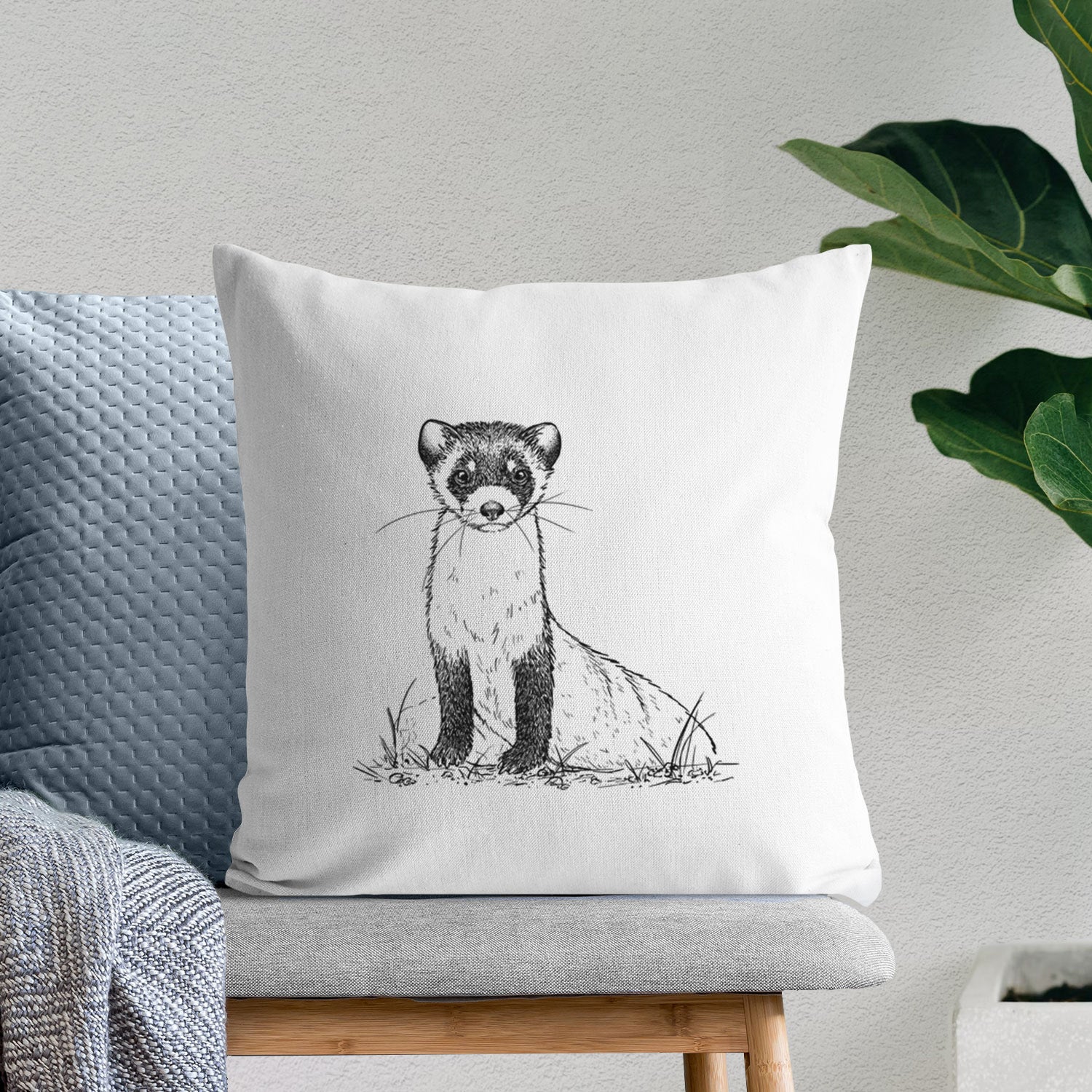White pillow cover printed with a detailed black ink illustration of a black-footed ferret sitting on the ground, on a bench beside plants.