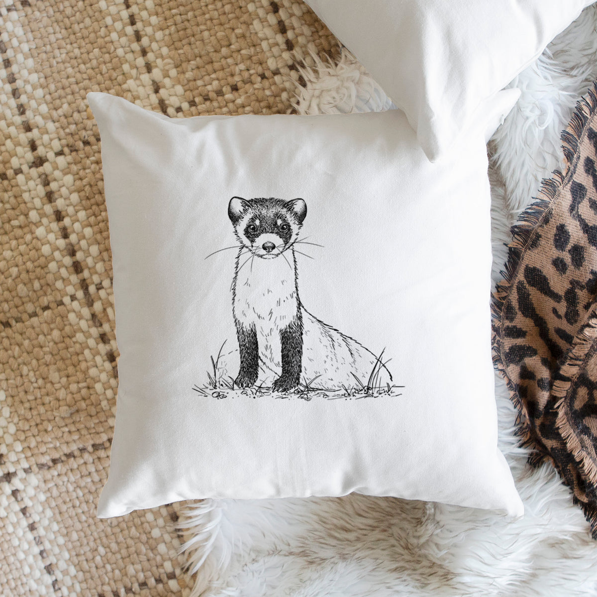 White pillow cover printed with a detailed black ink illustration of a black-footed ferret sitting on the ground, on a woven chair.
