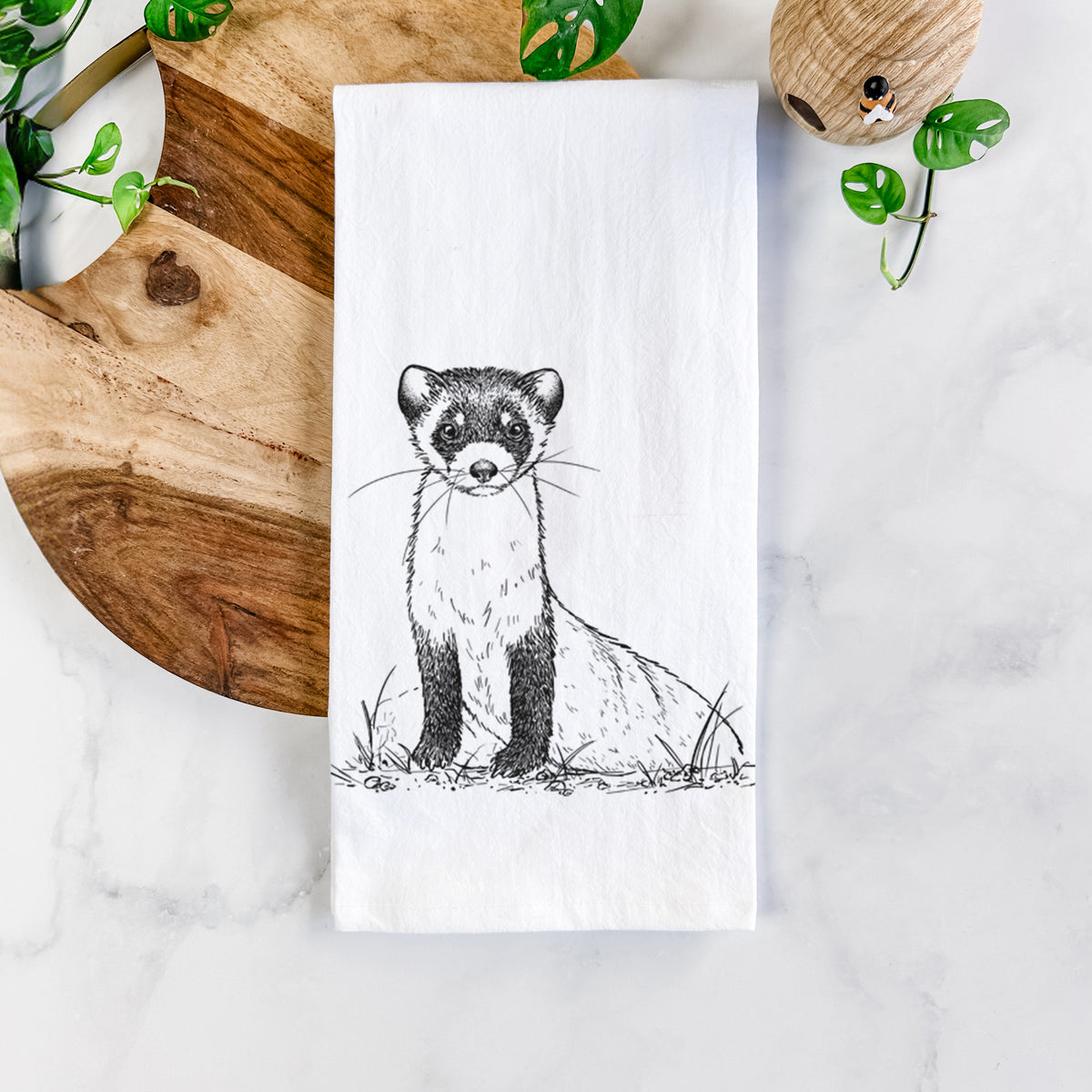 White cotton tea towel printed with a detailed black ink illustration of a black-footed ferret sitting on the ground, on a marble counter beside a round wooden cutting board and greenery.