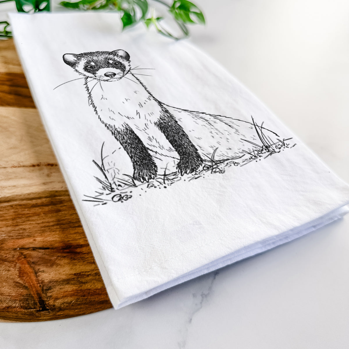 Close-up of a folded white tea towel on a wooden board, showing a detailed black ink illustration of a black-footed ferret sitting on the ground.