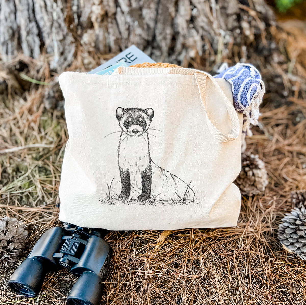 Canvas tote bag printed with a detailed black ink illustration of a black-footed ferret sitting on the ground, resting on dry grass beside a pair of binoculars.