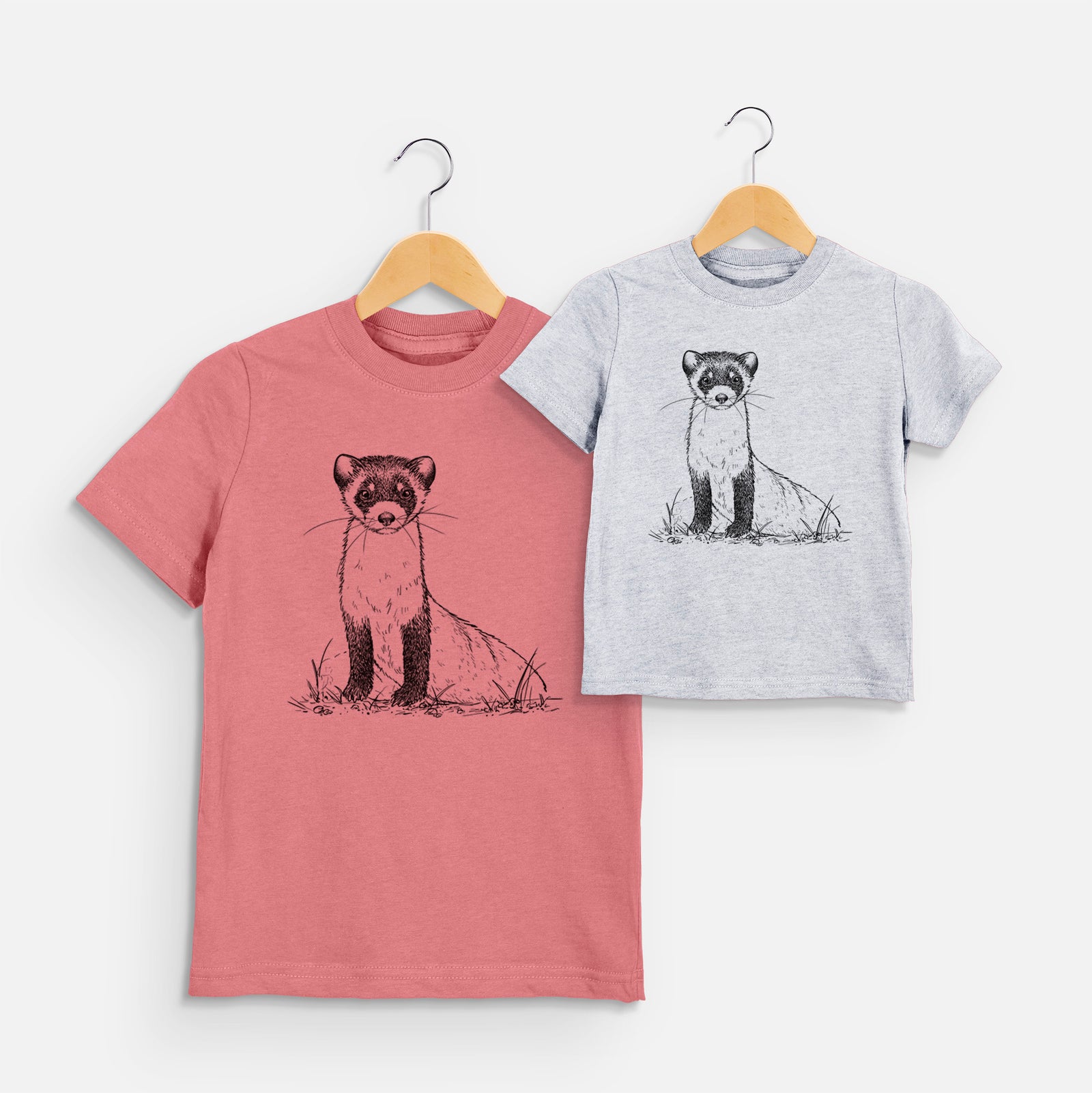 Two youth t-shirts in different colors on hangers, printed with a detailed black ink illustration of a black-footed ferret sitting on the ground.
