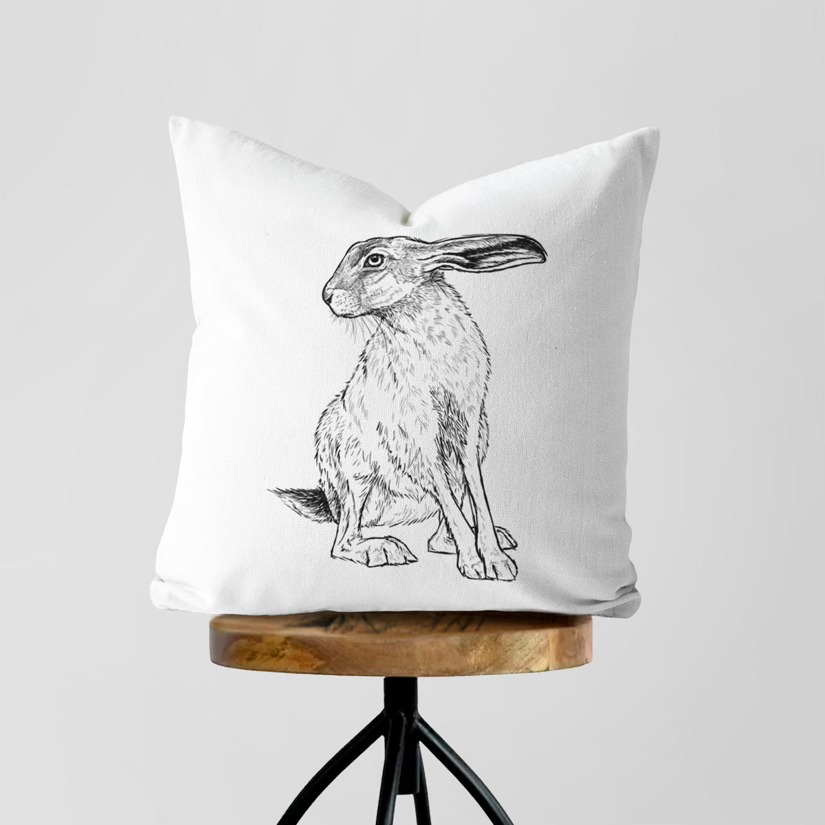 White pillow cover printed with a detailed black ink illustration of a black-tailed jackrabbit sitting with long upright ears, on a wooden stool.