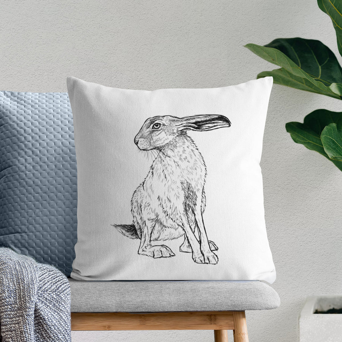White pillow cover printed with a detailed black ink illustration of a black-tailed jackrabbit sitting with long upright ears, on a bench beside plants.