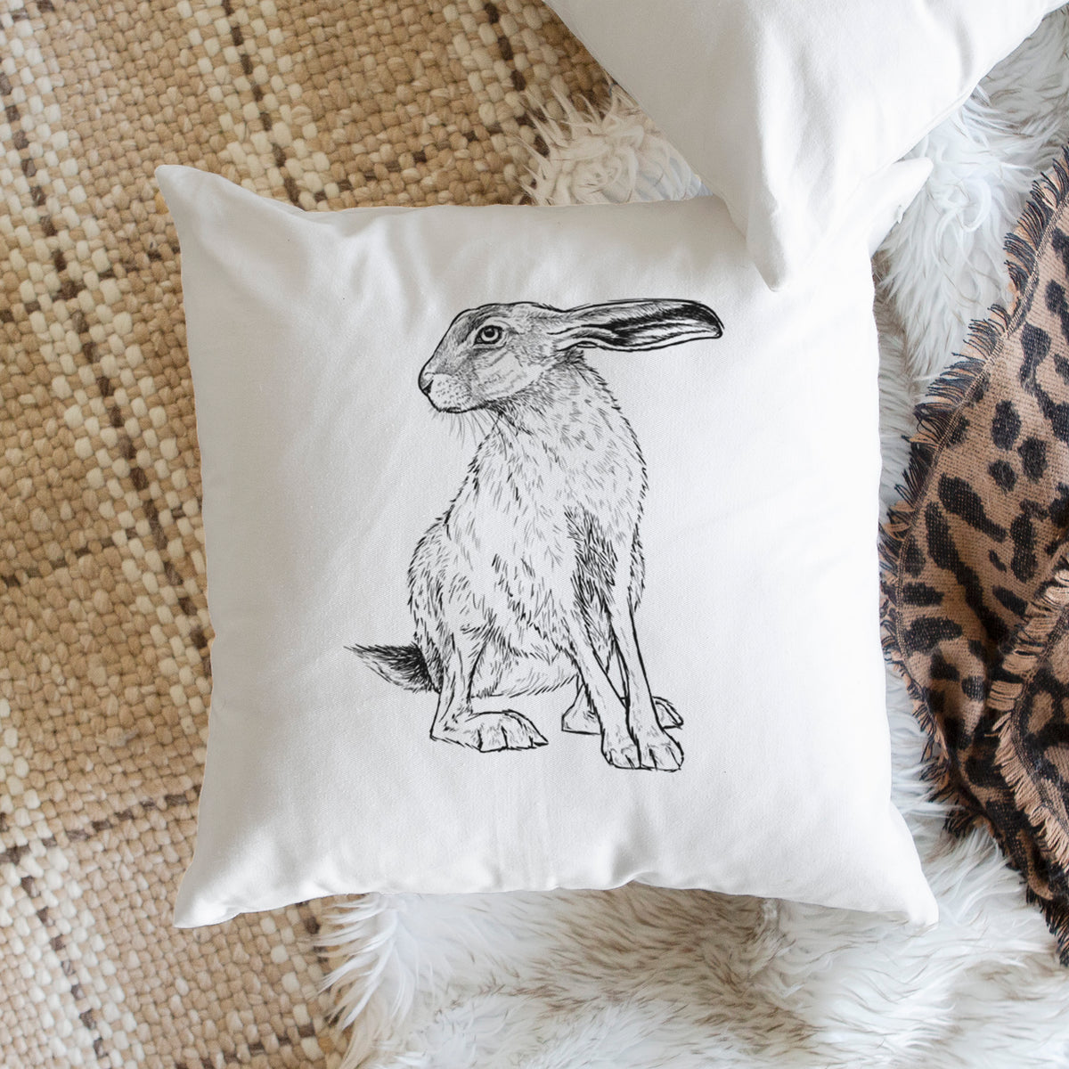 White pillow cover printed with a detailed black ink illustration of a black-tailed jackrabbit sitting with long upright ears, on a woven chair.