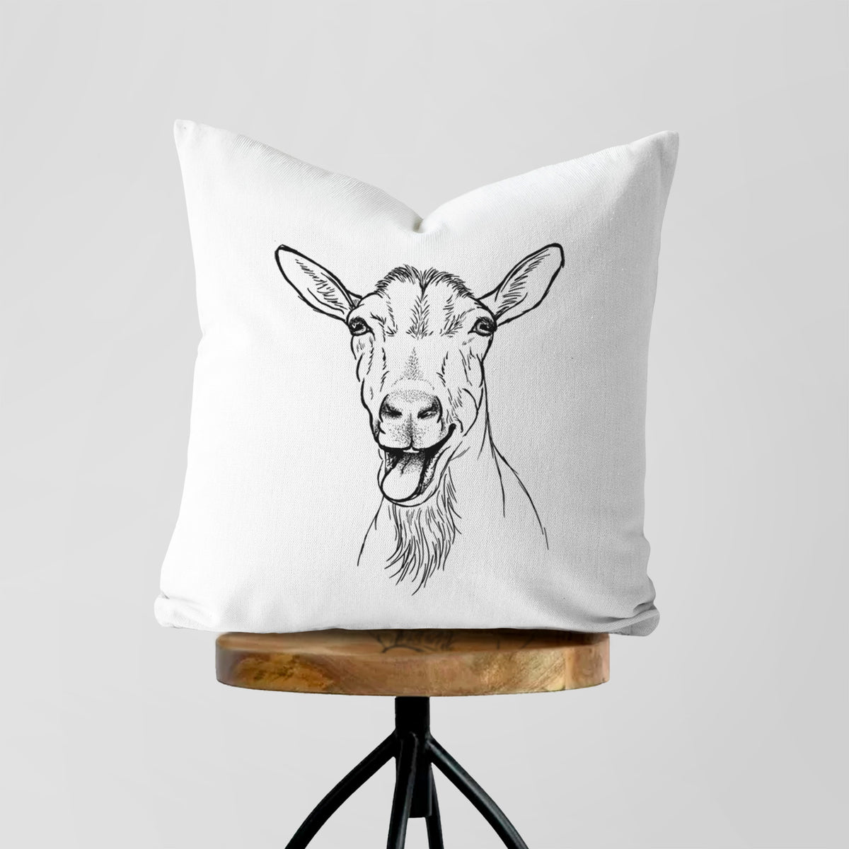 White pillow cover printed with a black ink illustration of a goat's head with its mouth open mid-bleat and its tongue out, on a wooden stool.