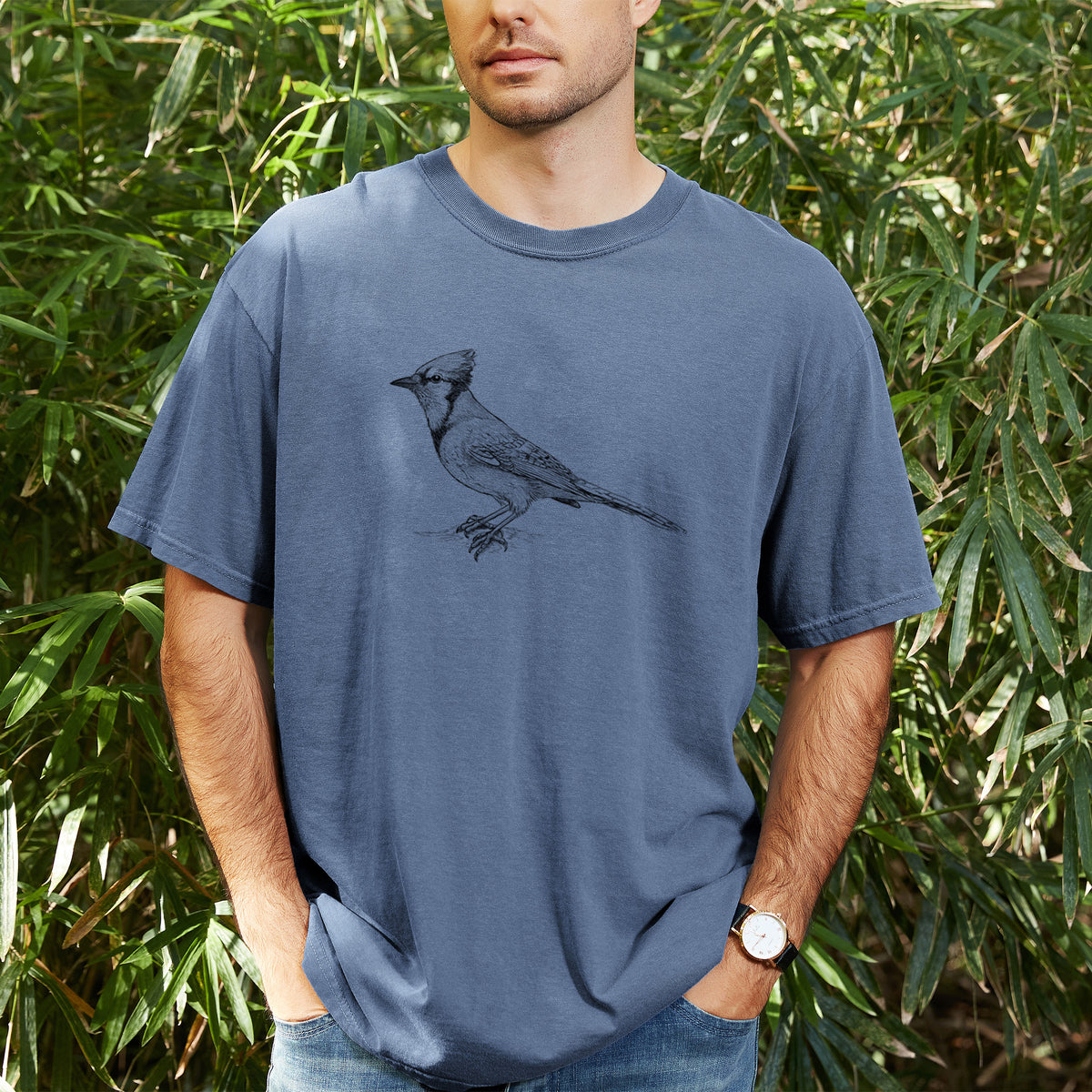 Man wearing a Comfort Colors t-shirt printed with a detailed black ink sketch of a blue jay perched with its crest raised, outdoors.