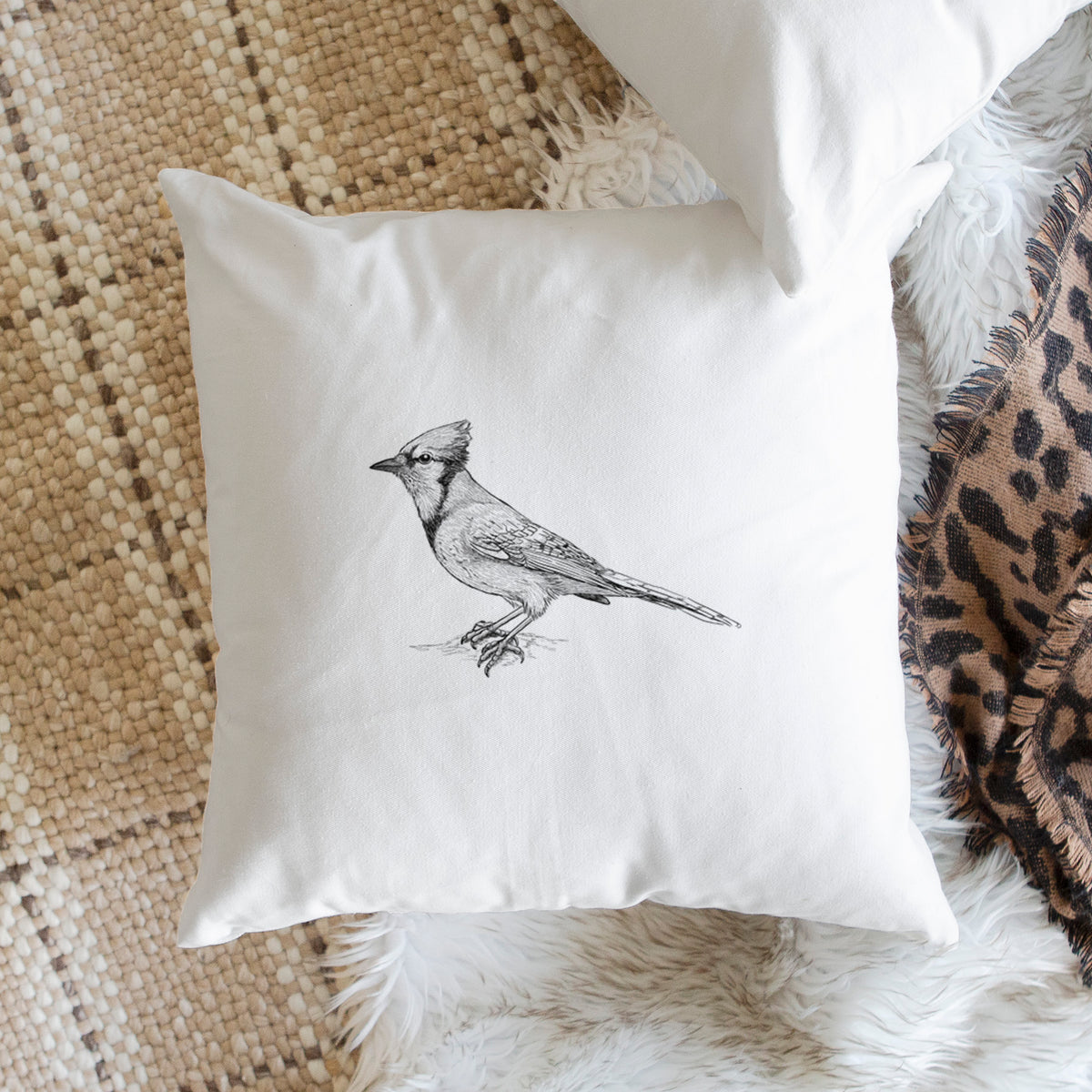 White pillow cover printed with a detailed black ink sketch of a blue jay perched with its crest raised, on a woven chair.