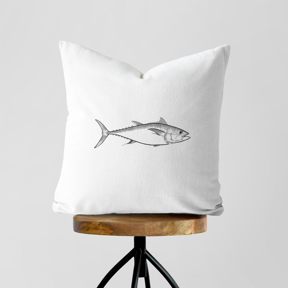 White pillow cover printed with a black ink line drawing of a bluefin tuna swimming, on a wooden stool.