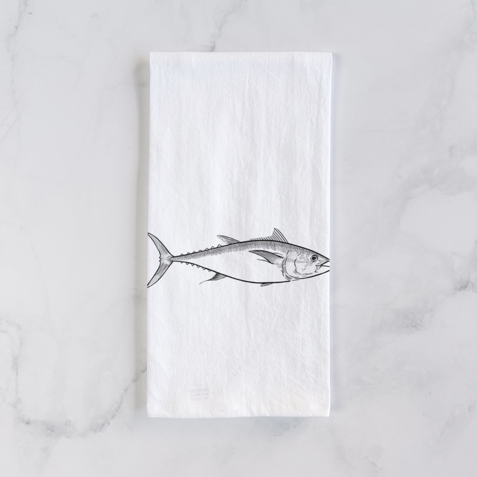White cotton tea towel printed with a black ink line drawing of a bluefin tuna swimming, on a marble counter beside a round wooden cutting board and greenery.