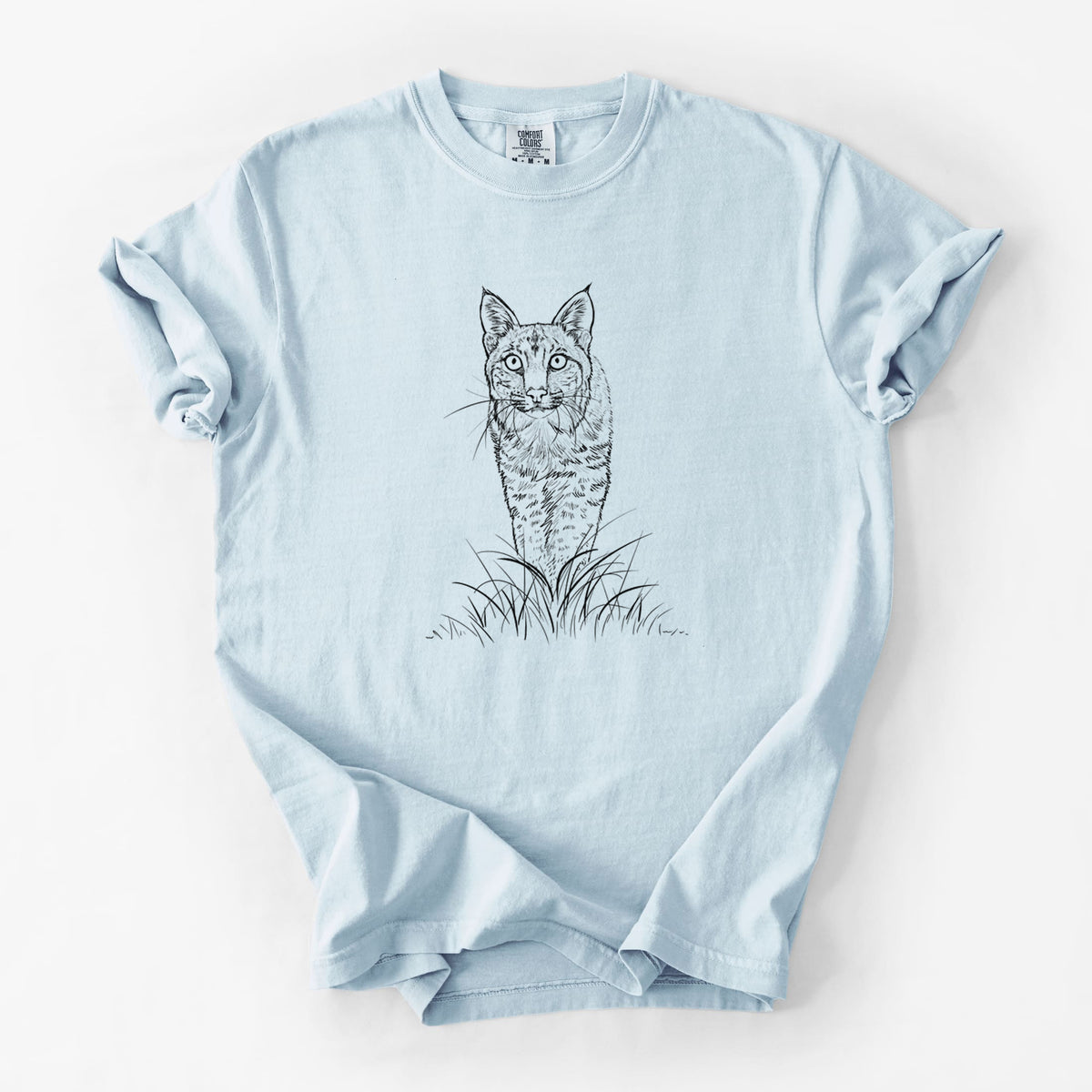 Chambray blue Comfort Colors t-shirt laid flat, printed with a black ink illustration of a bobcat standing in tall grass, facing forward.
