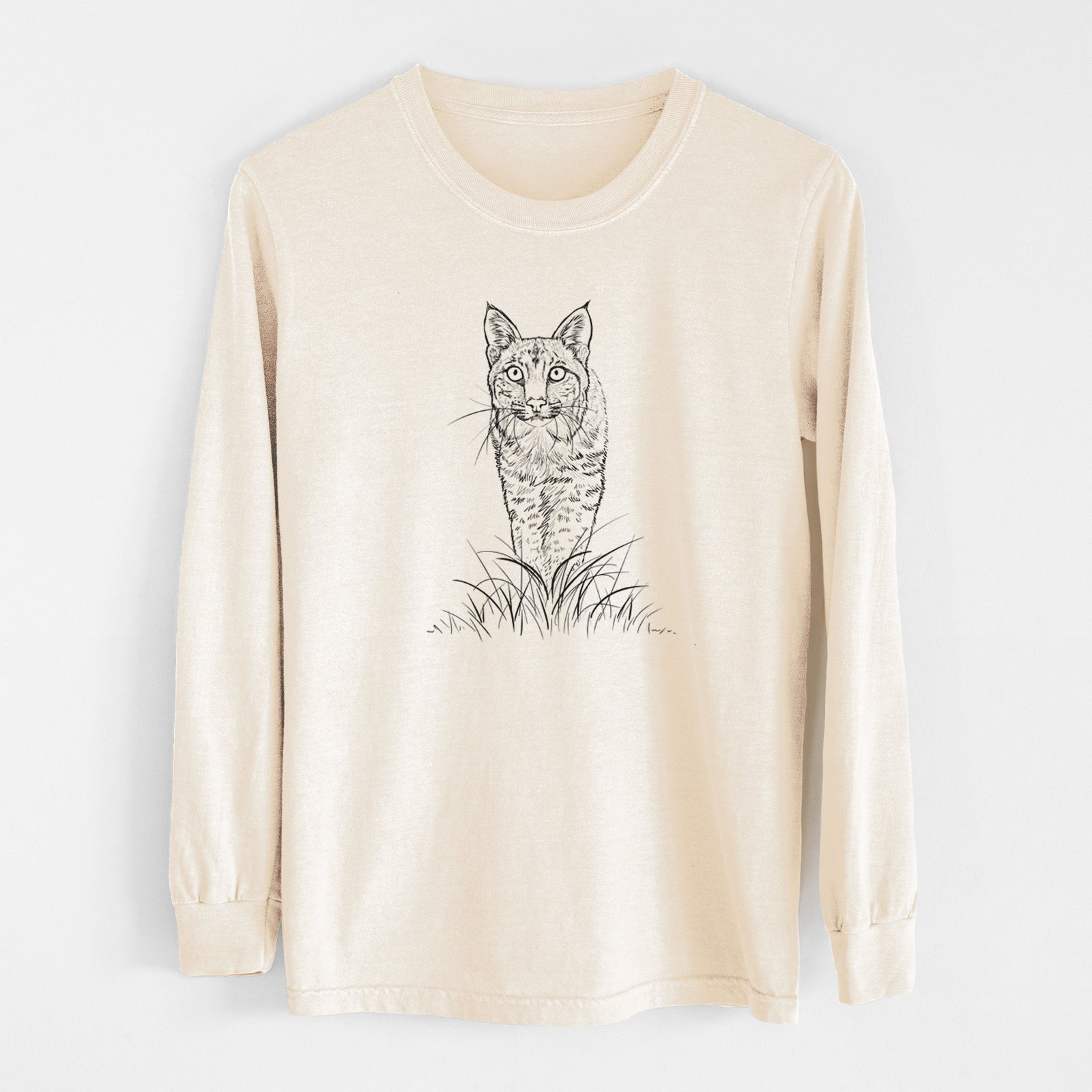 Ivory Comfort Colors long sleeve shirt laid flat, printed with a black ink illustration of a bobcat standing in tall grass, facing forward.