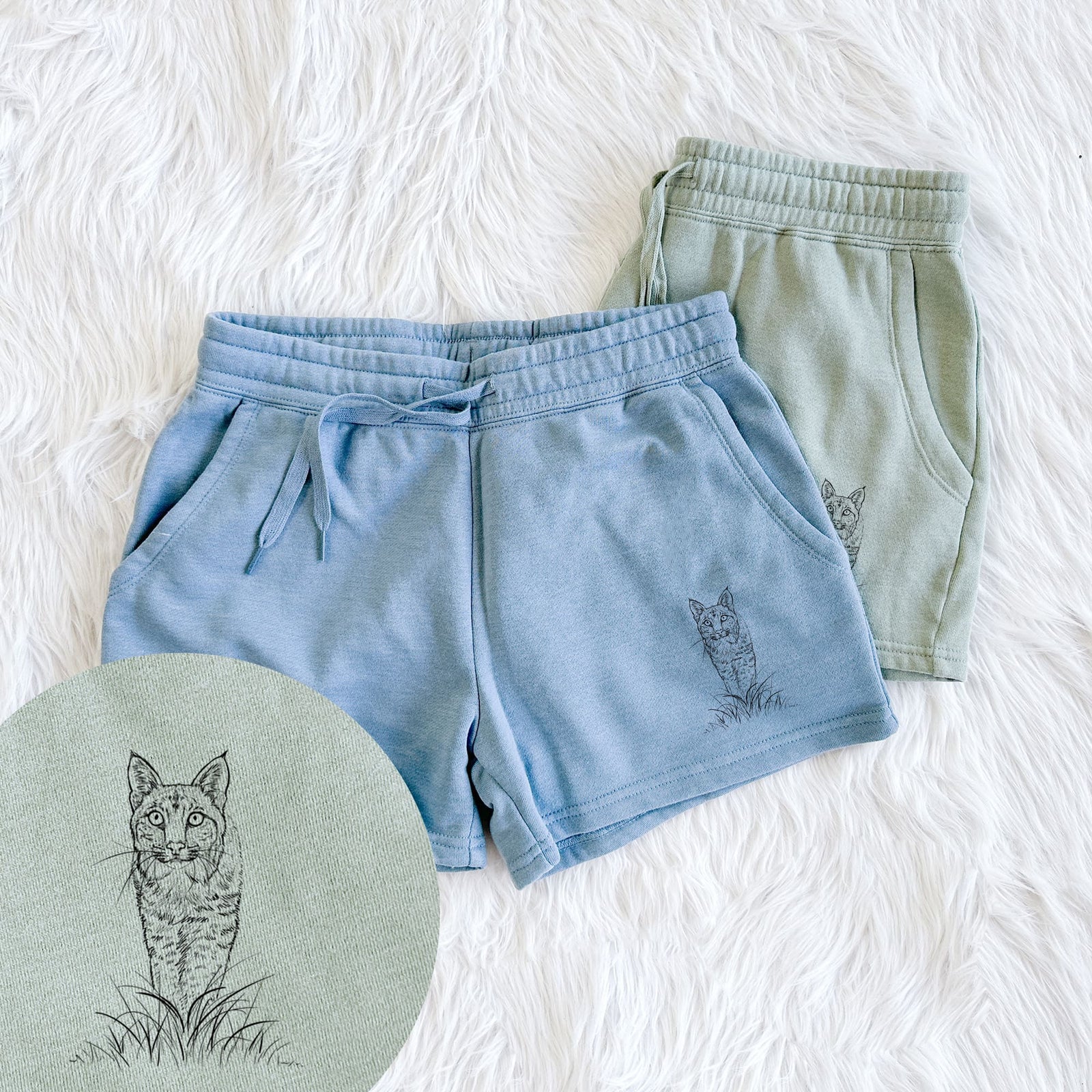 Two pairs of Cali Wave shorts in different colors printed with a black ink illustration of a bobcat standing in tall grass, facing forward, with a close-up of the print.