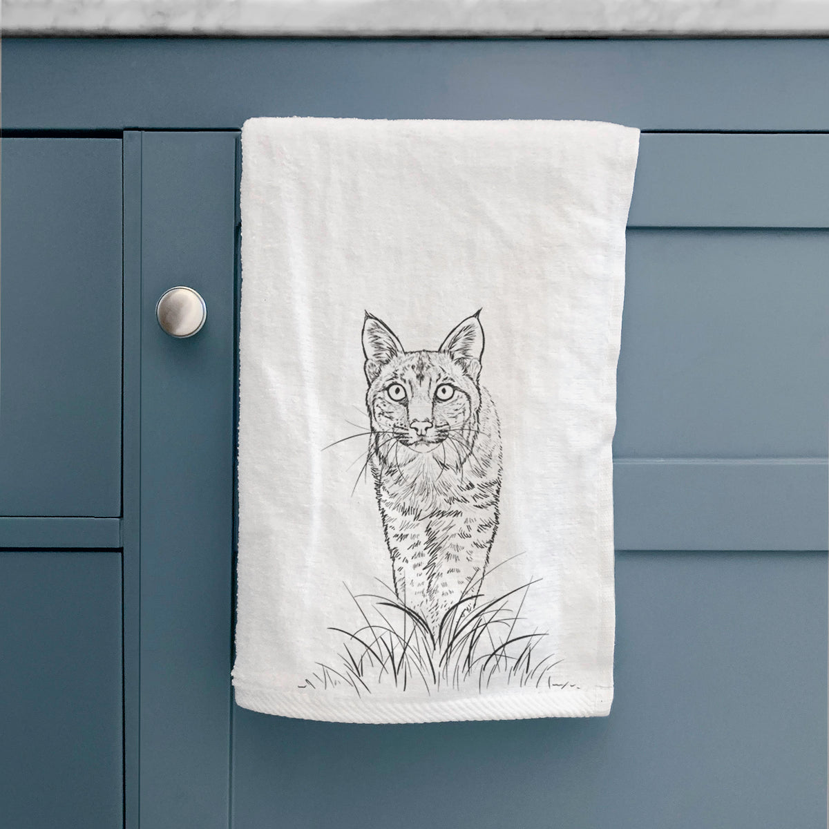 White hand towel printed with a black ink illustration of a bobcat standing in tall grass, facing forward, hanging on a cabinet door.