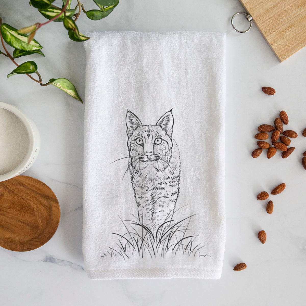 White hand towel printed with a black ink illustration of a bobcat standing in tall grass, facing forward.