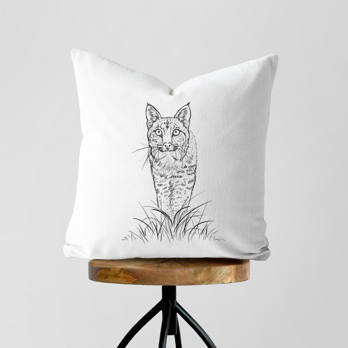 White pillow cover printed with a black ink illustration of a bobcat standing in tall grass, facing forward, on a wooden stool.