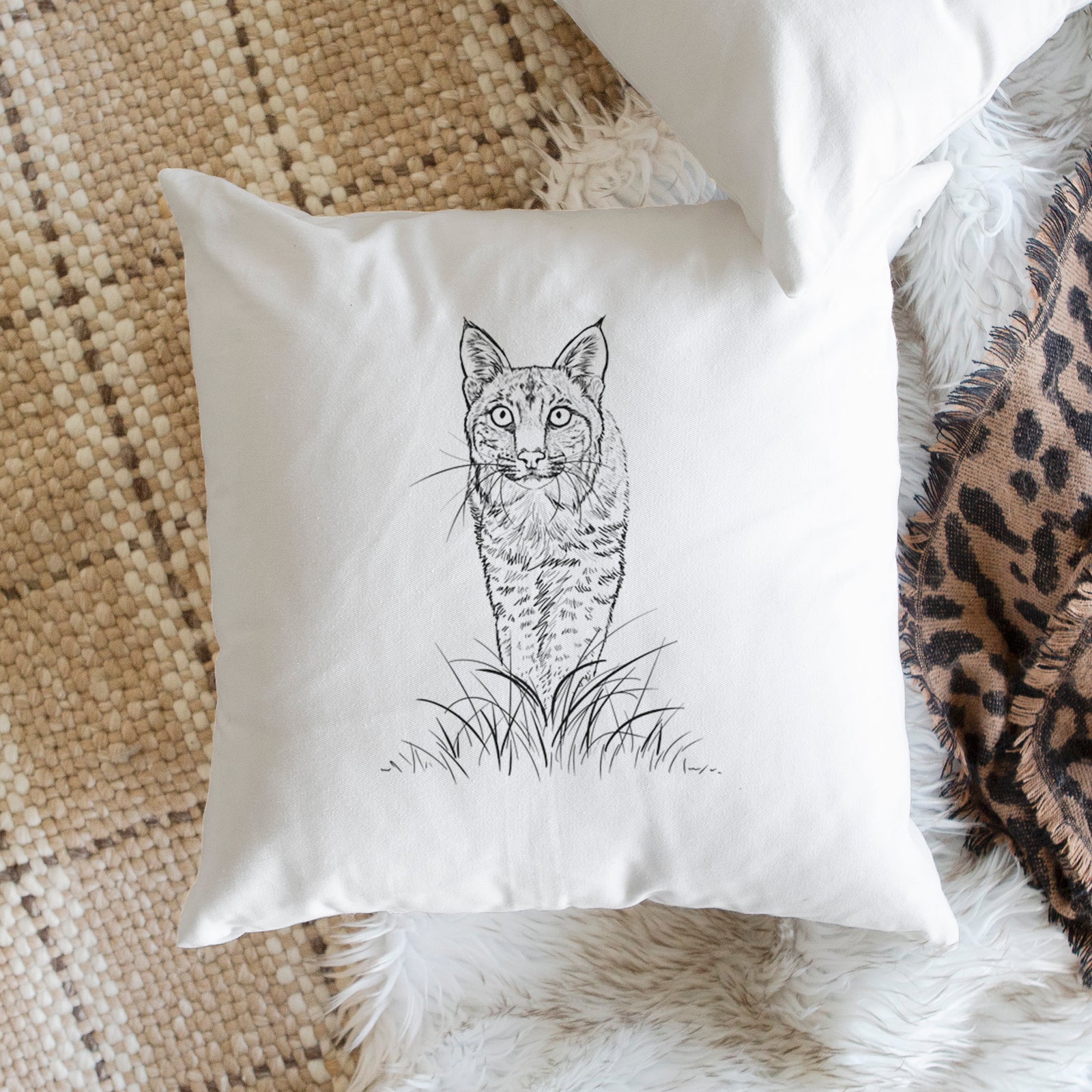 White pillow cover printed with a black ink illustration of a bobcat standing in tall grass, facing forward, on a bench beside plants.
