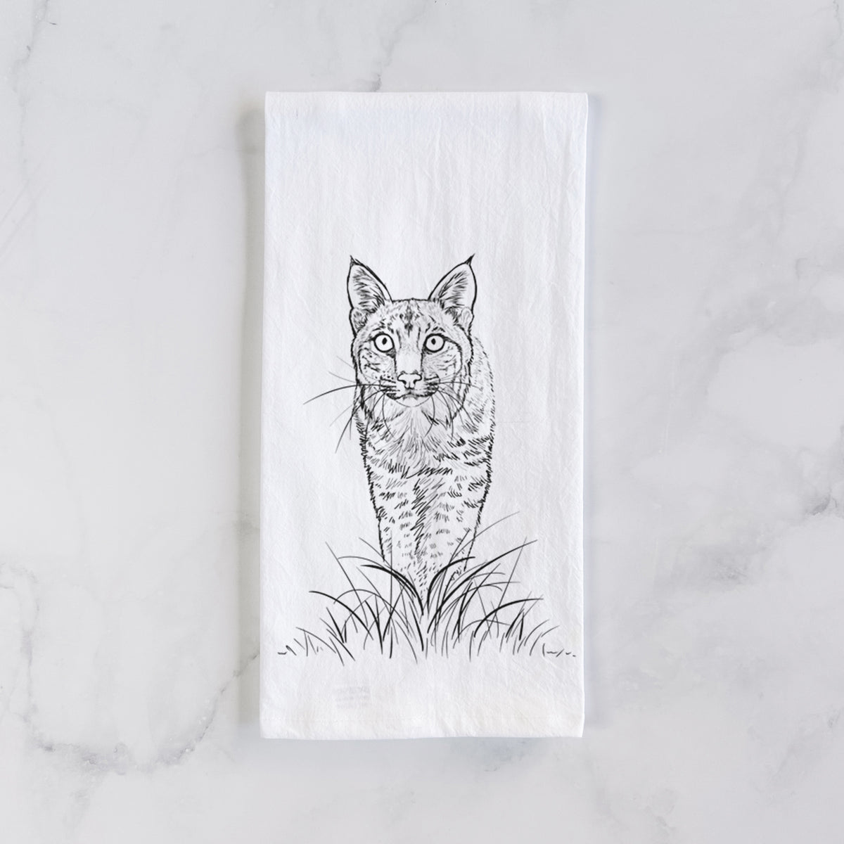 White cotton tea towel laid flat on marble, printed with a black ink illustration of a bobcat standing in tall grass, facing forward.