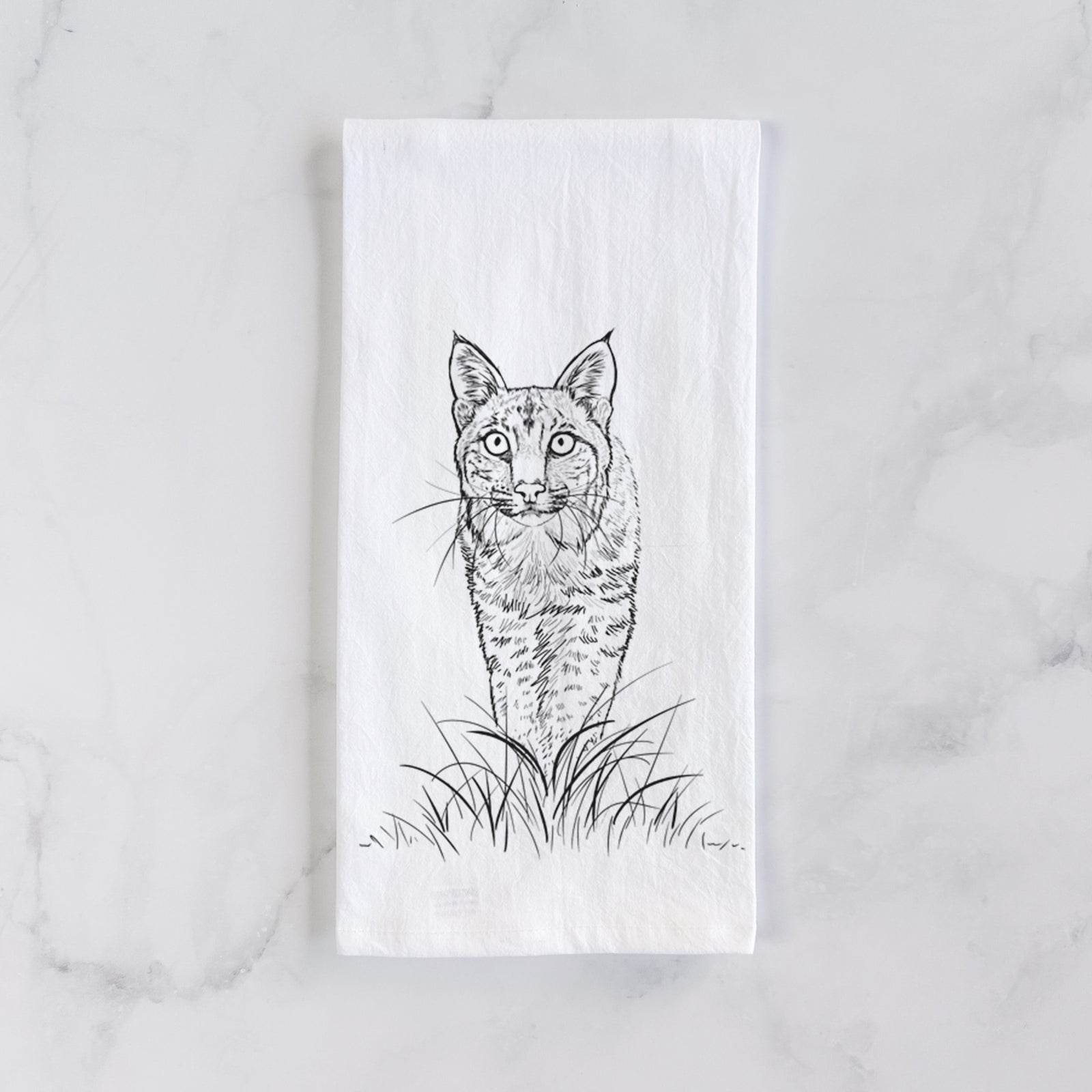 White cotton tea towel printed with a black ink illustration of a bobcat standing in tall grass, facing forward, on a marble counter beside a round wooden cutting board and greenery.