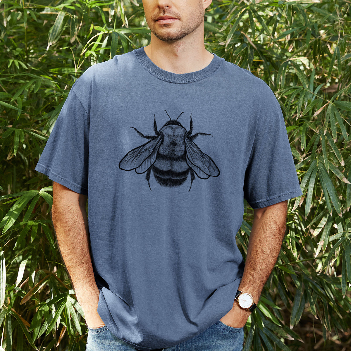 Man wearing a Comfort Colors t-shirt printed with a detailed black ink illustration of a rusty patched bumble bee with its wings spread, seen from above, outdoors.