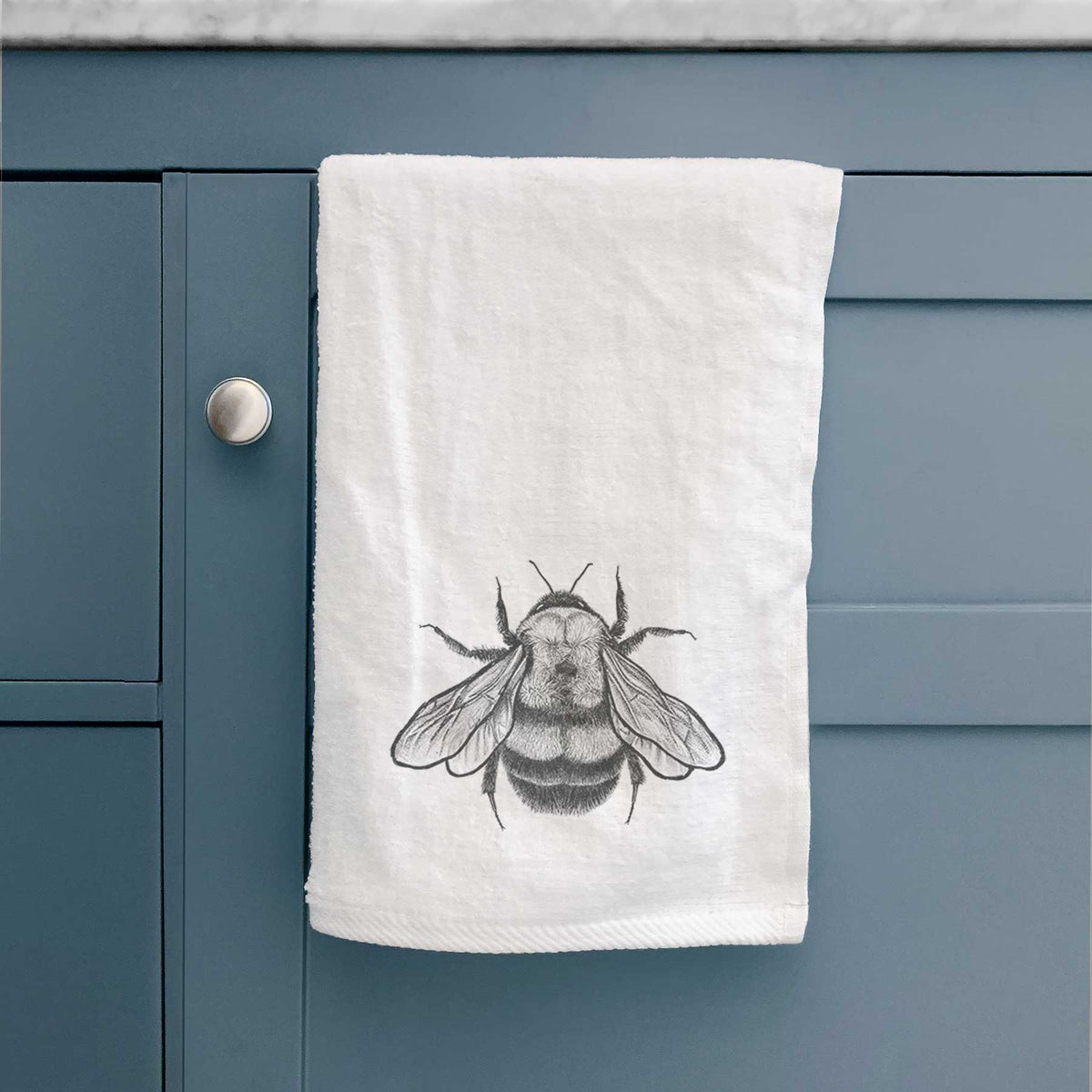 White hand towel printed with a detailed black ink illustration of a rusty patched bumble bee with its wings spread, seen from above, hanging on a cabinet door.