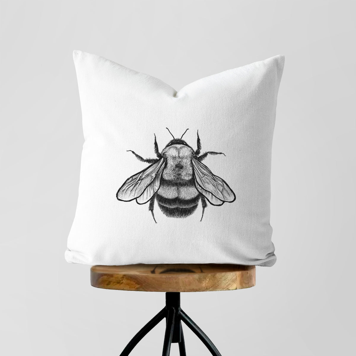White pillow cover printed with a detailed black ink illustration of a rusty patched bumble bee with its wings spread, seen from above, on a wooden stool.