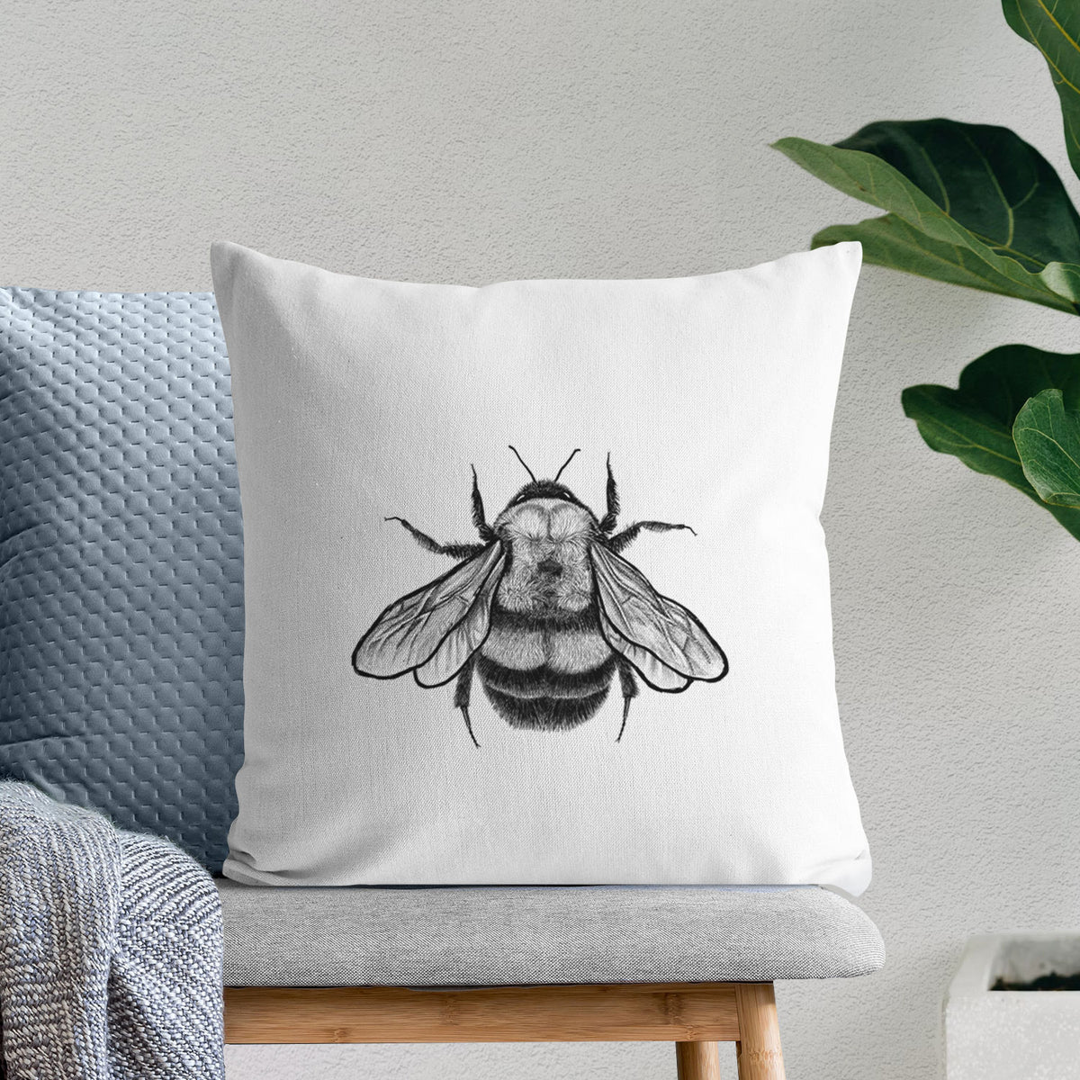 White pillow cover printed with a detailed black ink illustration of a rusty patched bumble bee with its wings spread, seen from above, on a bench beside plants.