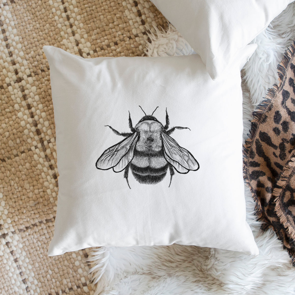 White pillow cover printed with a detailed black ink illustration of a rusty patched bumble bee with its wings spread, seen from above, on a woven chair.