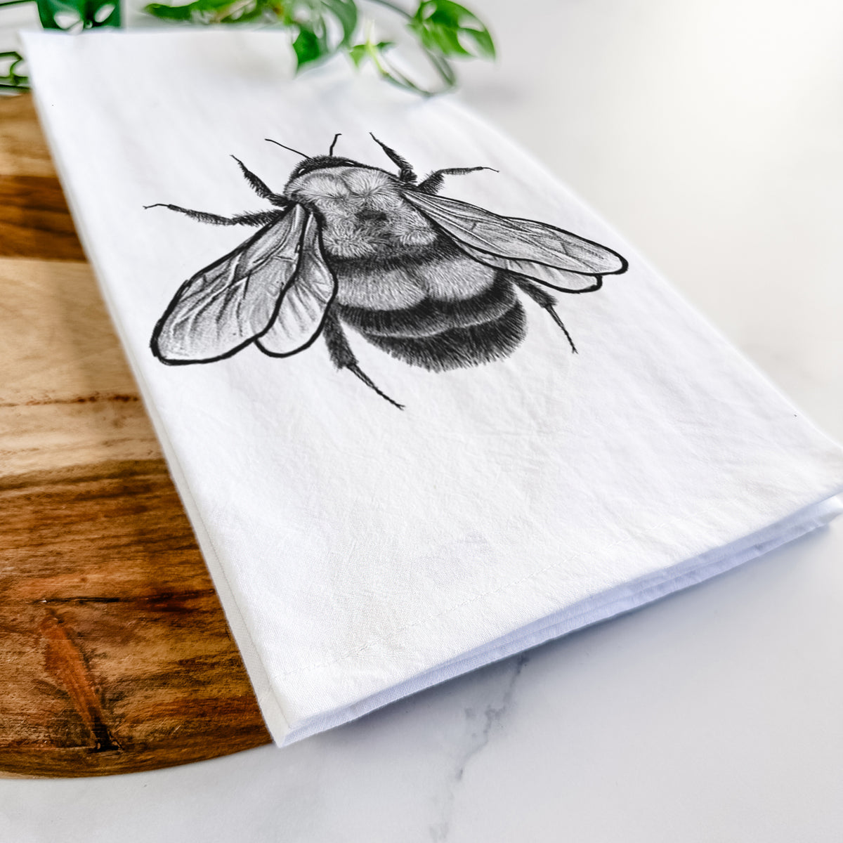 Close-up of a folded white tea towel on a wooden board, showing a detailed black ink illustration of a rusty patched bumble bee with its wings spread, seen from above.