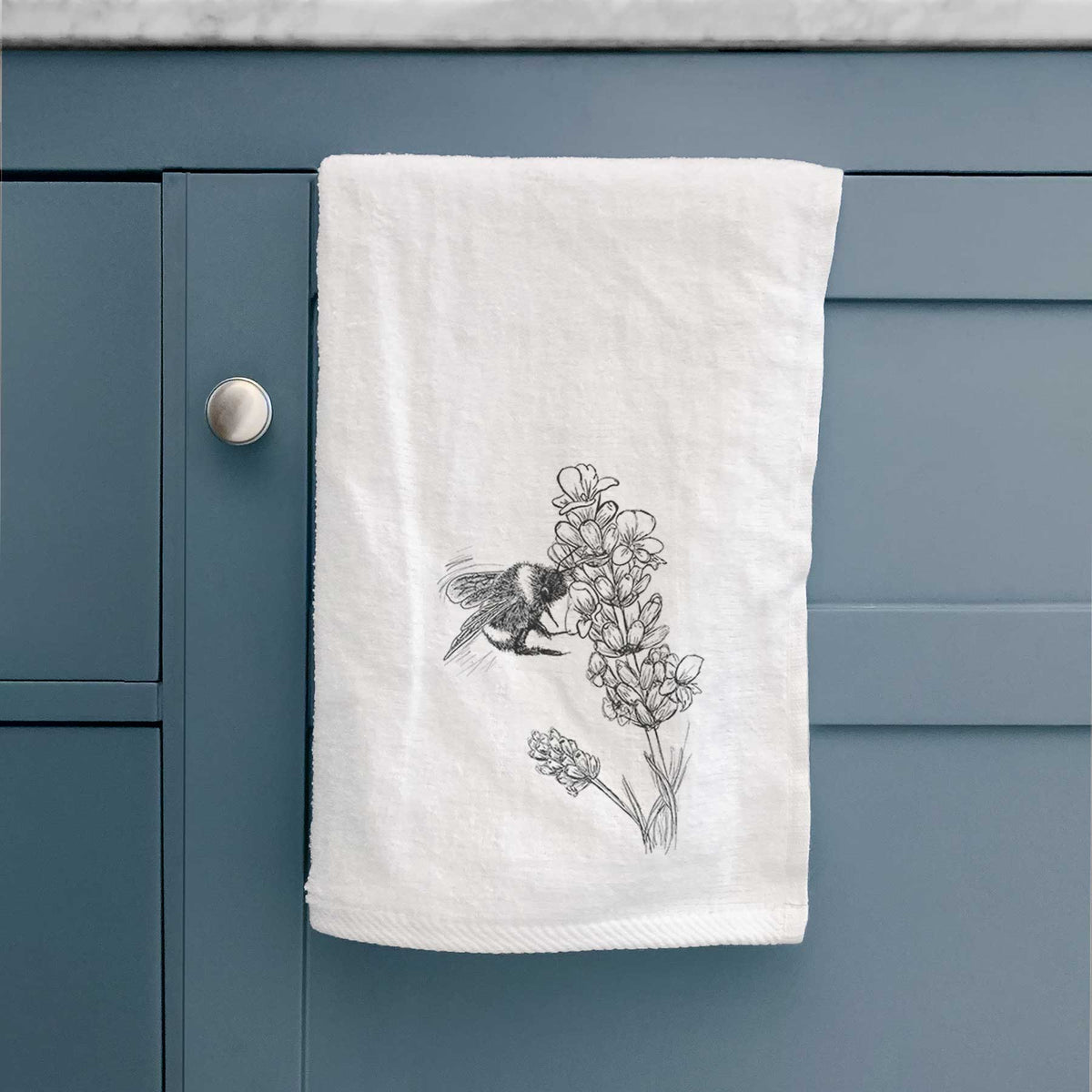 White hand towel printed with a black ink illustration of a bumble bee visiting a sprig of lavender, hanging on a cabinet door.