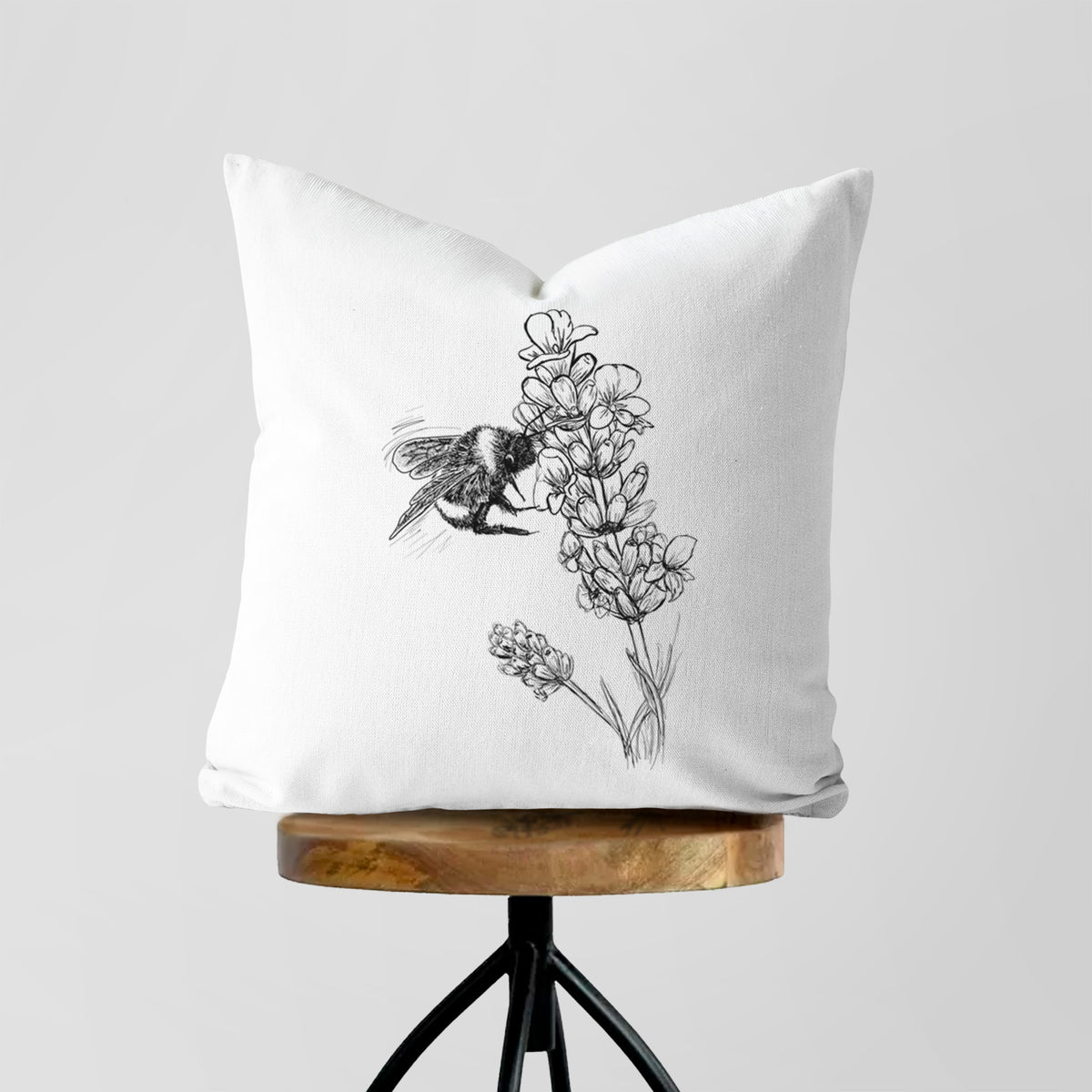 White pillow cover printed with a black ink illustration of a bumble bee visiting a sprig of lavender, on a wooden stool.