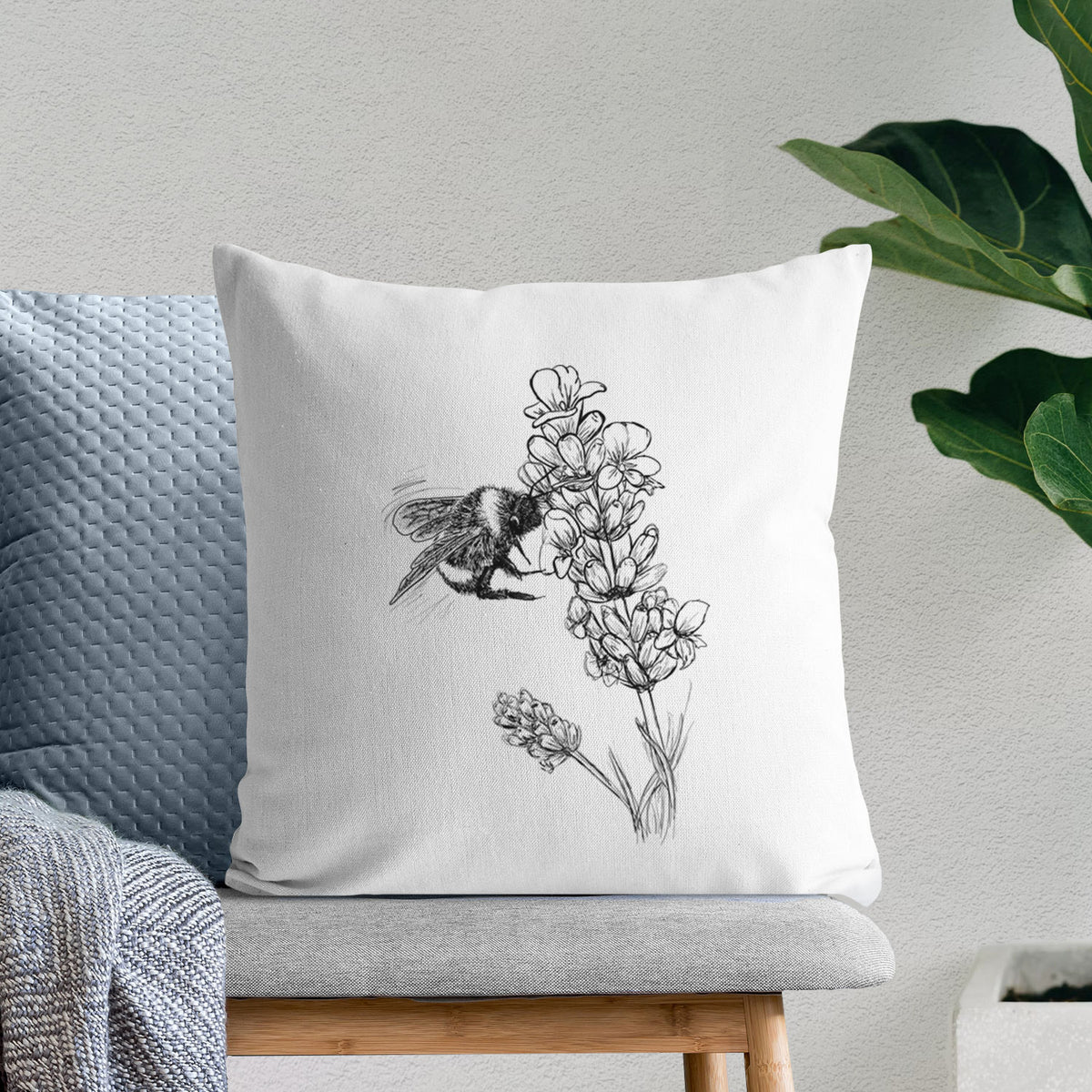 White pillow cover printed with a black ink illustration of a bumble bee visiting a sprig of lavender, on a bench beside plants.