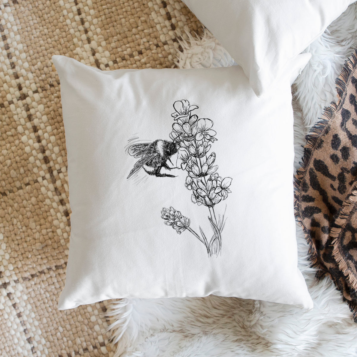 White pillow cover printed with a black ink illustration of a bumble bee visiting a sprig of lavender, on a woven chair.