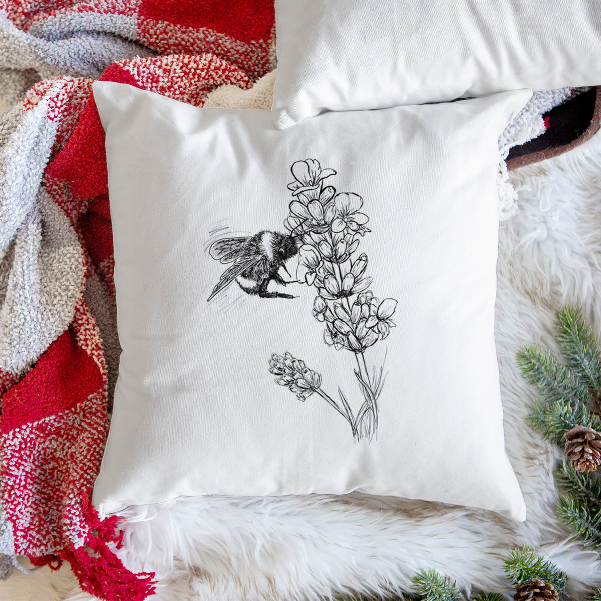 White pillow cover printed with a black ink illustration of a bumble bee visiting a sprig of lavender, on a woven chair.