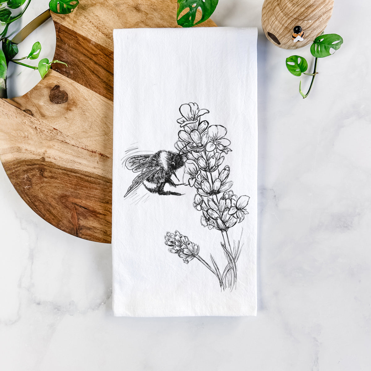 White cotton tea towel printed with a black ink illustration of a bumble bee visiting a sprig of lavender, on a marble counter beside a round wooden cutting board and greenery.