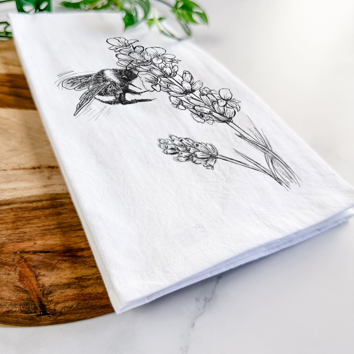 Close-up of a folded white tea towel on a wooden board, showing a black ink illustration of a bumble bee visiting a sprig of lavender.