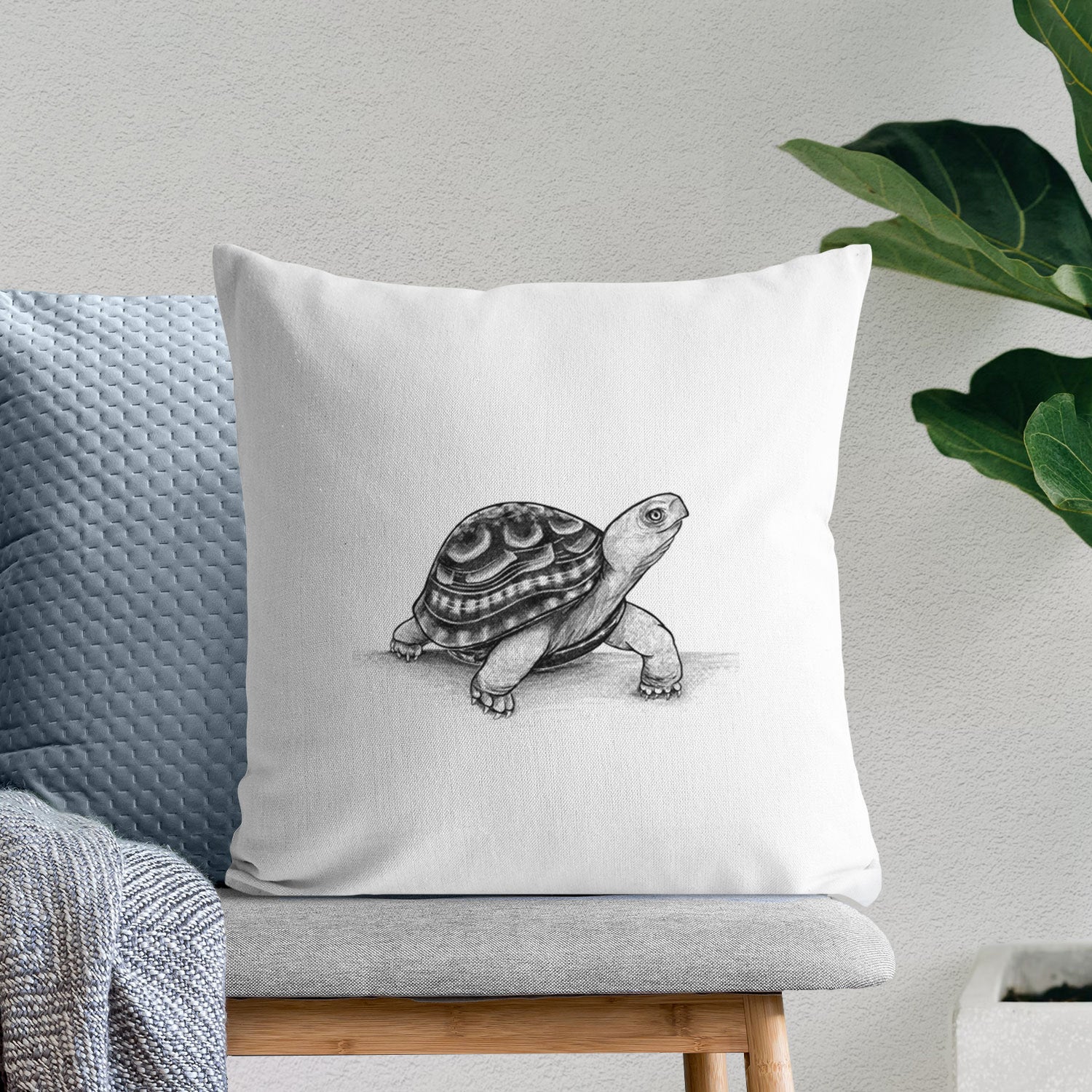 White pillow cover printed with a detailed black ink illustration of an eastern box turtle walking, on a bench beside plants.